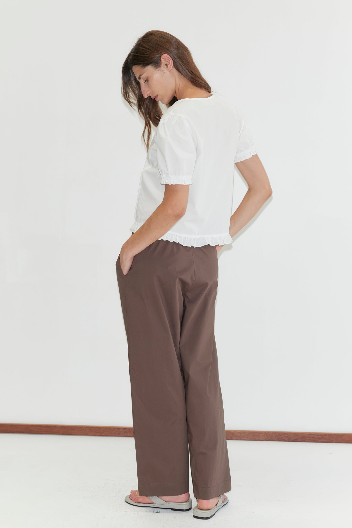 A woman stands against a plain white background, looking down with one hand in her brown wide-leg pants pocket. She wears the Pintuck Top - White from Deiji Studios and light sandals.