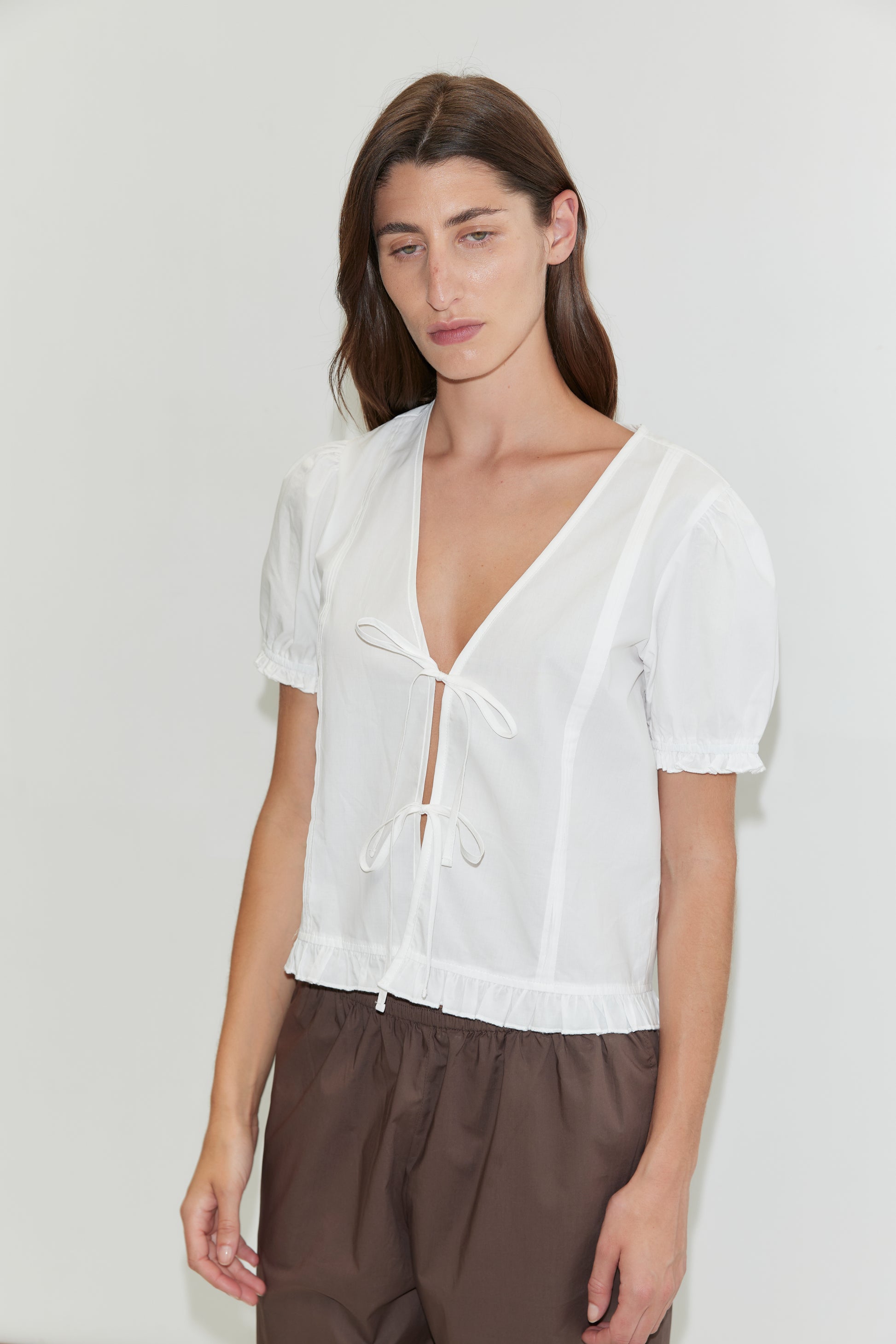 A person with long brown hair wears the Pintuck Top - White by Deiji Studios, featuring short sleeves, a deep V-neck, and front tie, paired with loose brown pants, standing against a plain white background.