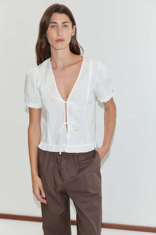 A person with long brown hair wears the Deiji Studios Pintuck Top in white, featuring short sleeves and front ties, paired with brown pants. They stand against a plain white background with one hand in their pocket.