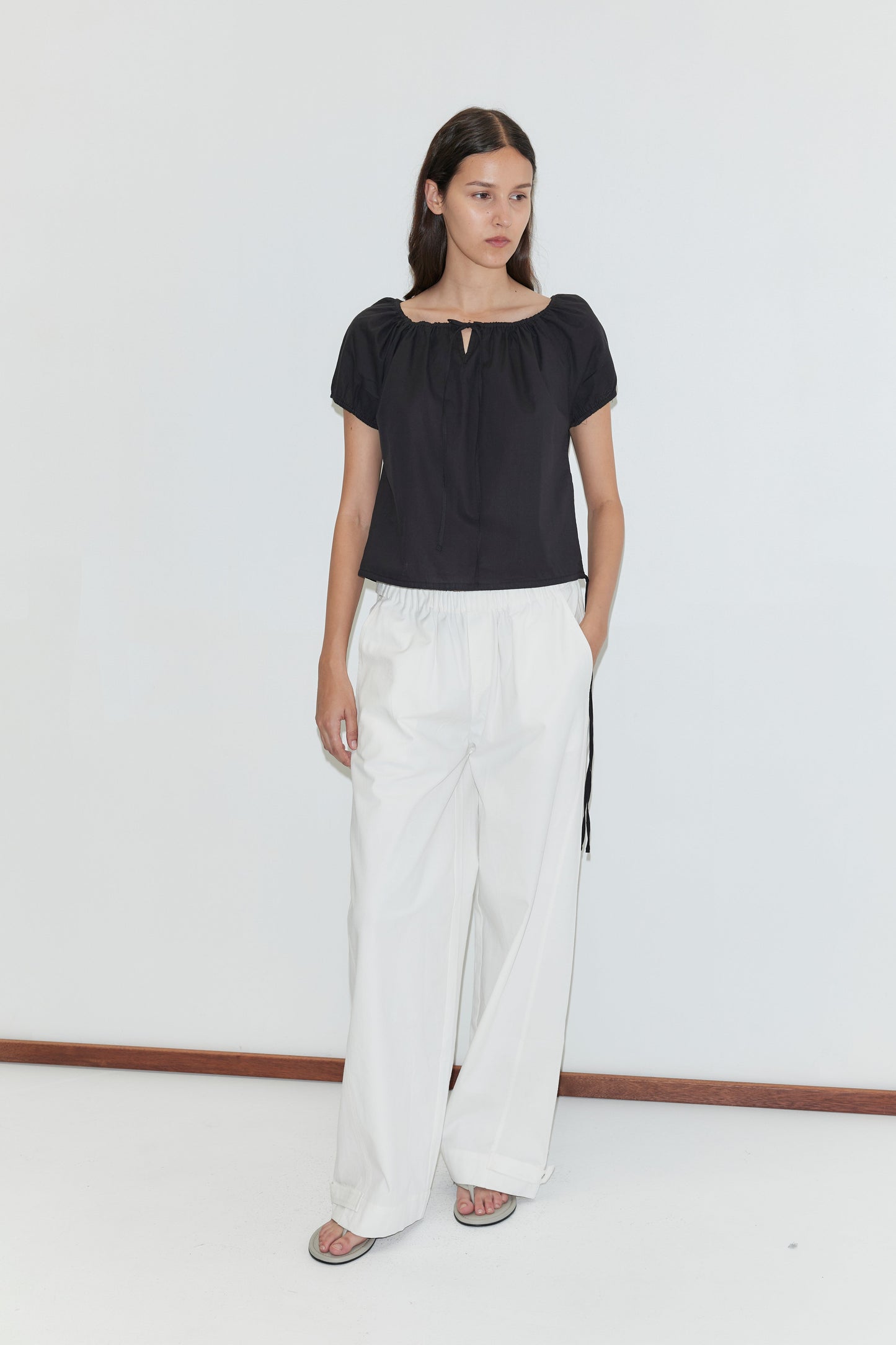 A woman with long dark hair and a neutral expression stands against a plain white wall, wearing Deiji Studios’ Tie Top - Black, wide-leg white pants, and sandals.