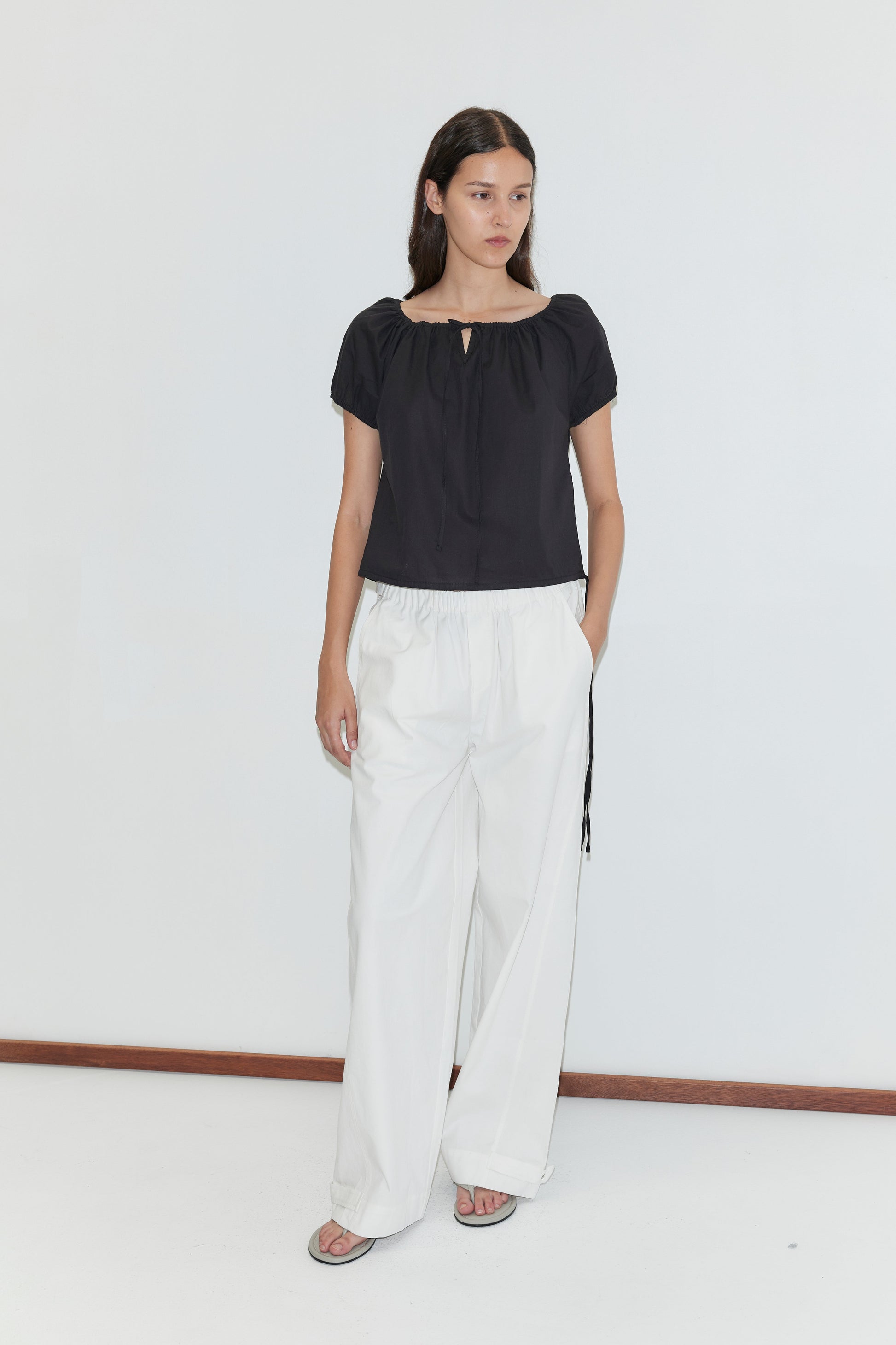 A woman with long dark hair and a neutral expression stands against a plain white wall, wearing Deiji Studios’ Tie Top - Black, wide-leg white pants, and sandals.