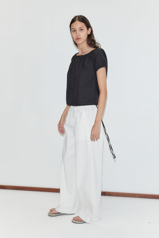 A woman with long brown hair and a neutral expression stands against a white background, wearing Deiji Studios' Tie Top in black, wide-legged white pants with a black side tie, and gray sandals.