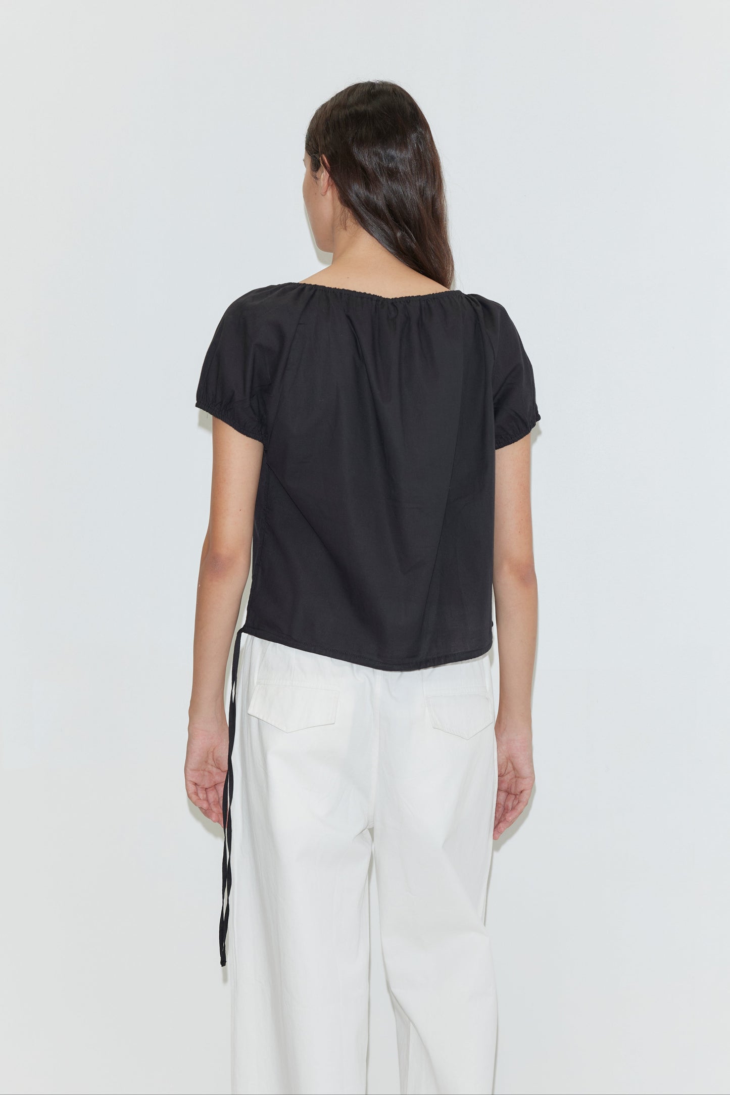 A person with long brown hair, seen from the back, wears the Deiji Studios Tie Top - Black and high-waisted white pants with large back pockets against a plain white background.