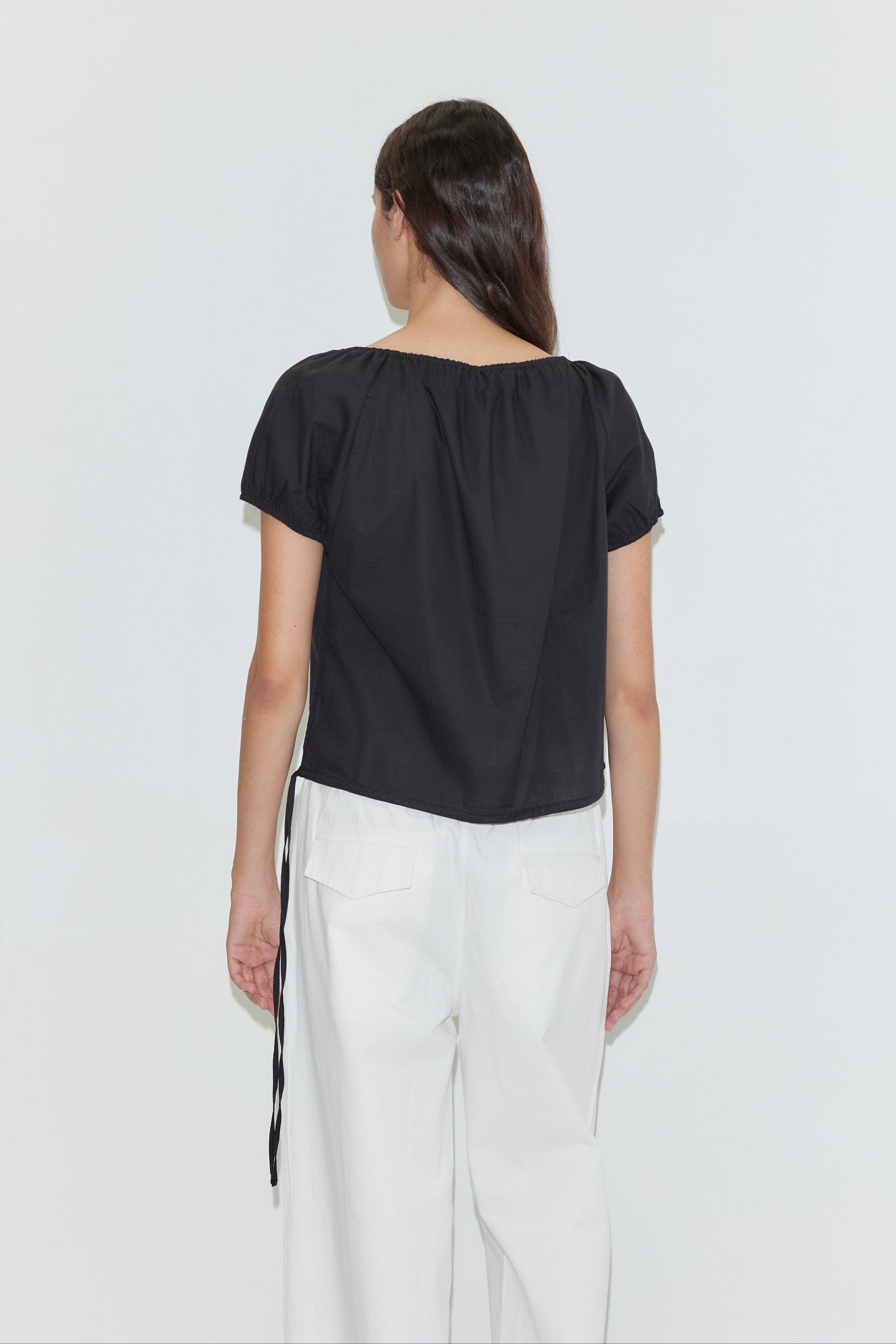 A person with long brown hair, seen from the back, wears the Deiji Studios Tie Top - Black and high-waisted white pants with large back pockets against a plain white background.