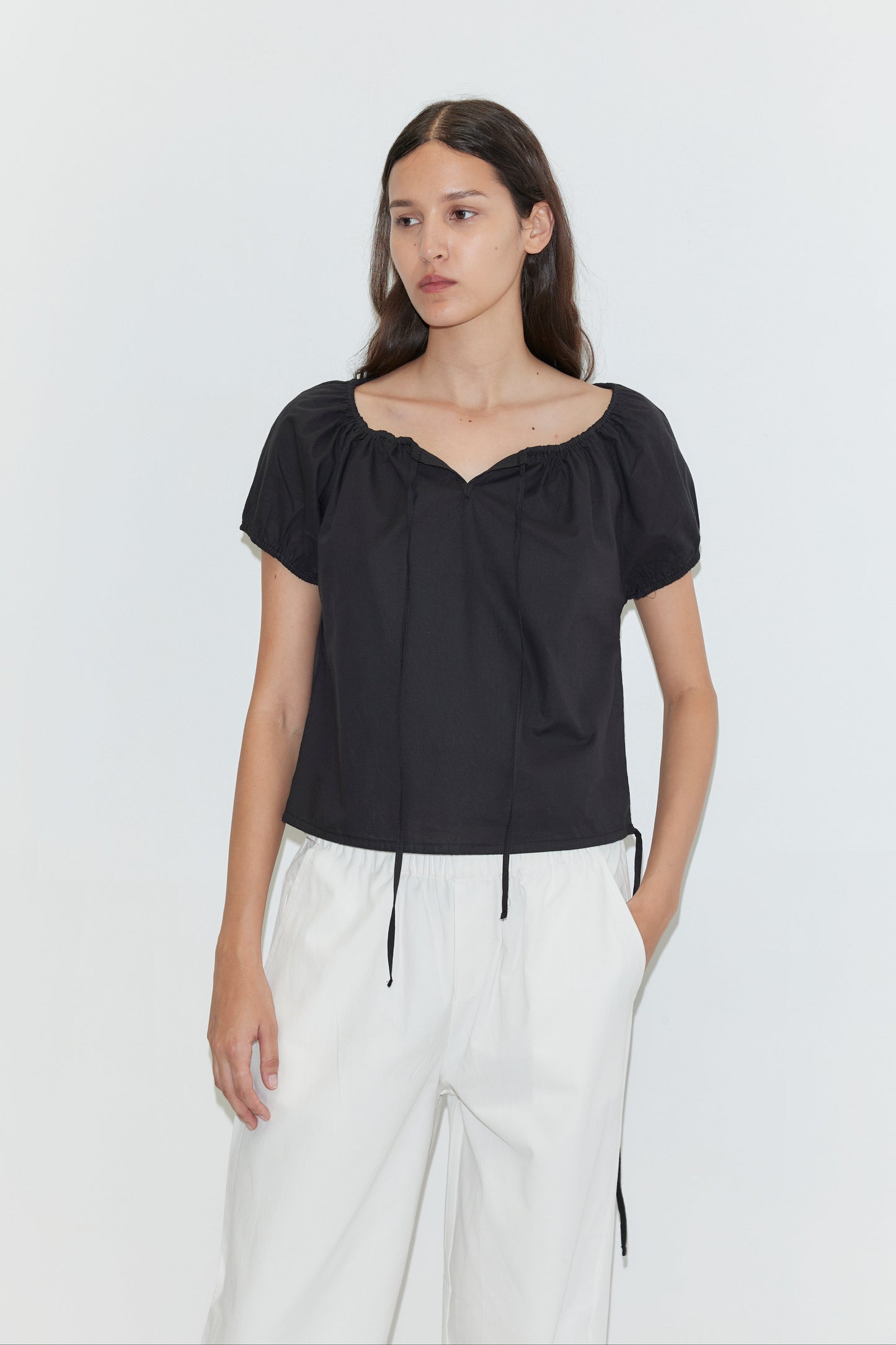 A person with long brown hair wears the Deiji Studios Tie Top - Black, a loose short-sleeve top with a gathered neckline, paired with white high-waisted pants against a plain white background.