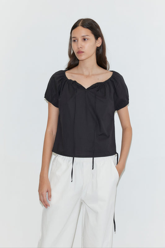 A person with long brown hair wears the Deiji Studios Tie Top - Black, a loose short-sleeve top with a gathered neckline, paired with white high-waisted pants against a plain white background.