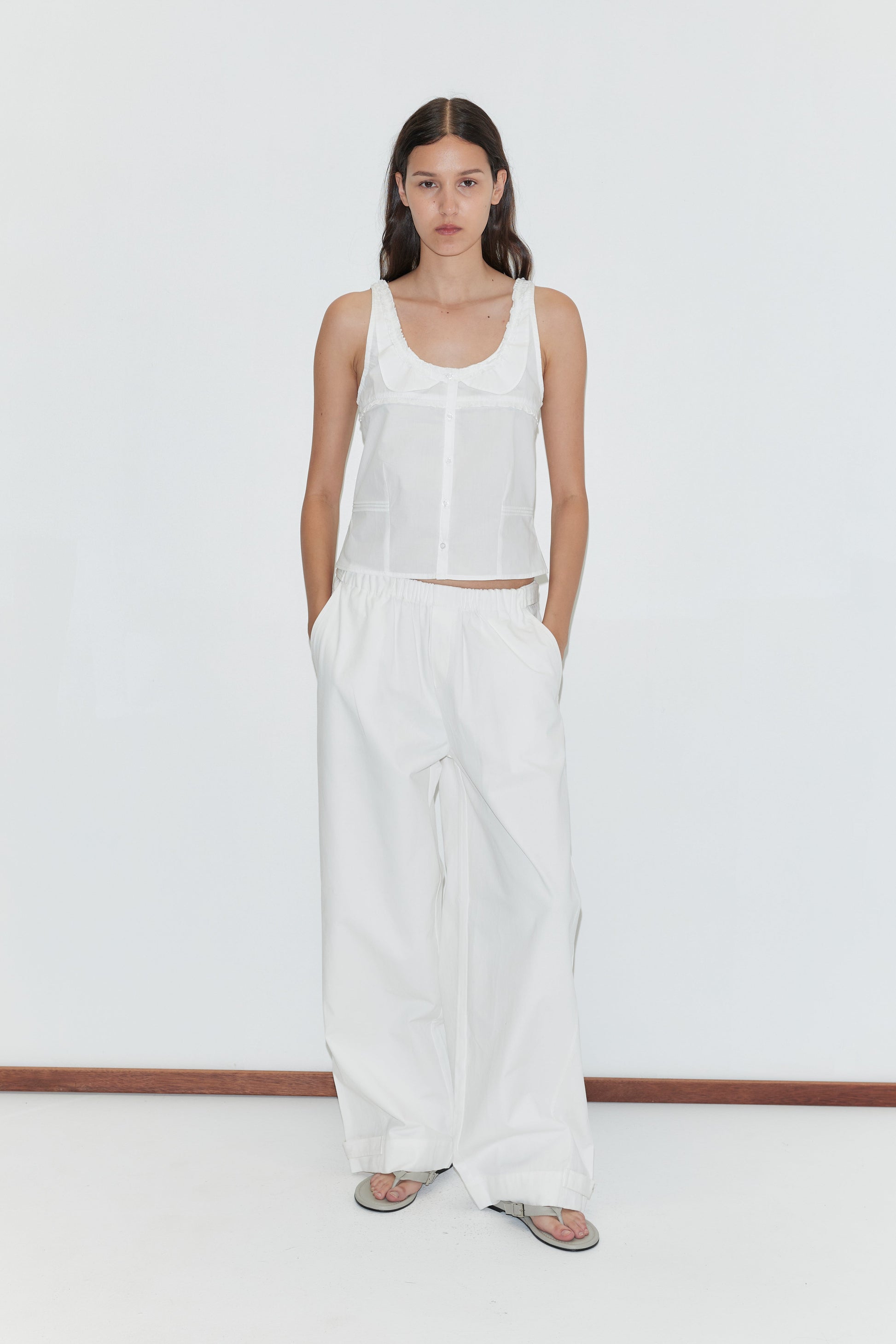 A woman with long brown hair stands against a white background wearing a sleeveless white button-up top, gray sandals, and the Snap Tab Pant - White by Deiji Studios, with her hands tucked casually in her pockets.