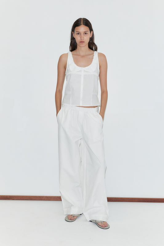 A woman with long brown hair stands against a white background wearing a sleeveless white button-up top, gray sandals, and the Snap Tab Pant - White by Deiji Studios, with her hands tucked casually in her pockets.