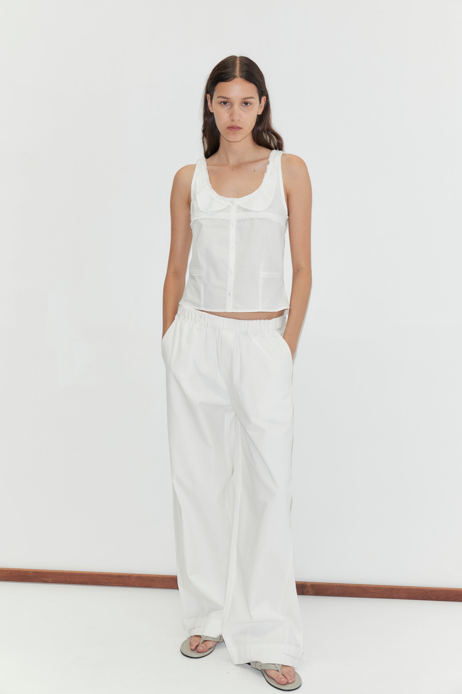 A woman with long, straight hair and a neutral expression stands against a plain white background, wearing the Frills Top - White by Deiji Studios, loose white pants with pockets, and gray flip-flops.