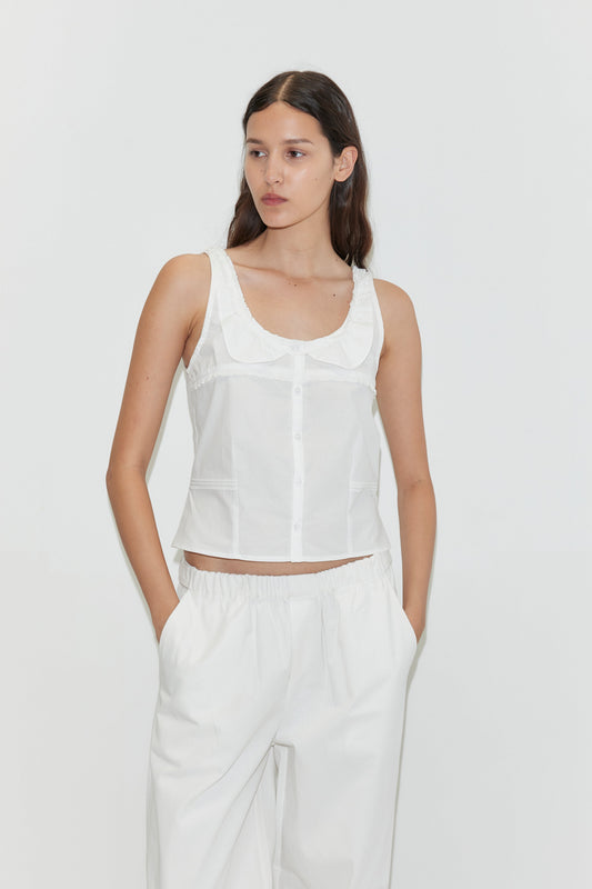 A woman with long brown hair wears the Frills Top - White by Deiji Studios, a sleeveless cotton poplin tank with a scoop collar, paired with loose white pants, standing against a plain white background with hands in her pockets.