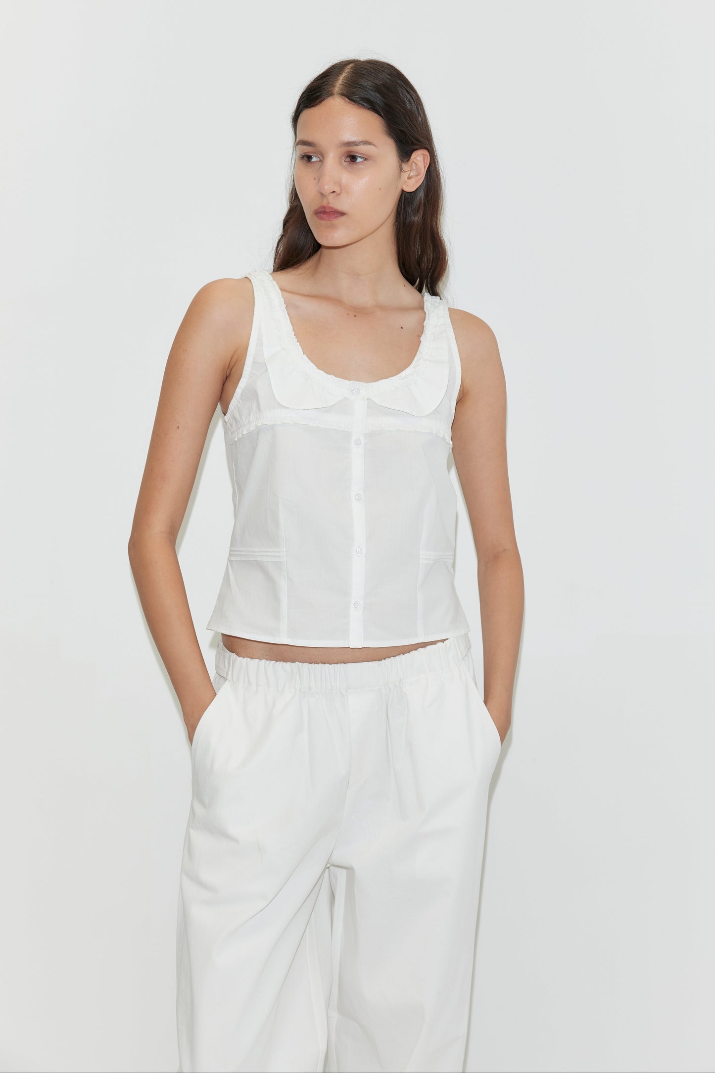 A woman with long dark hair wears a sleeveless white button-up top and loose-fitting Snap Tab Pant - White by Deiji Studios, standing against a plain white background with her hands in her pockets.