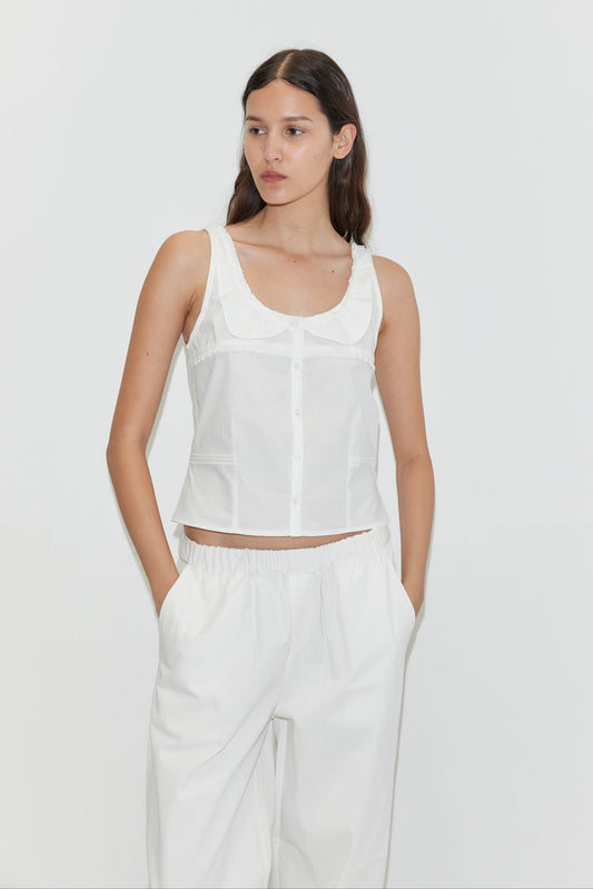 A woman with long dark hair wears a sleeveless white button-up top and loose-fitting Snap Tab Pant - White by Deiji Studios, standing against a plain white background with her hands in her pockets.