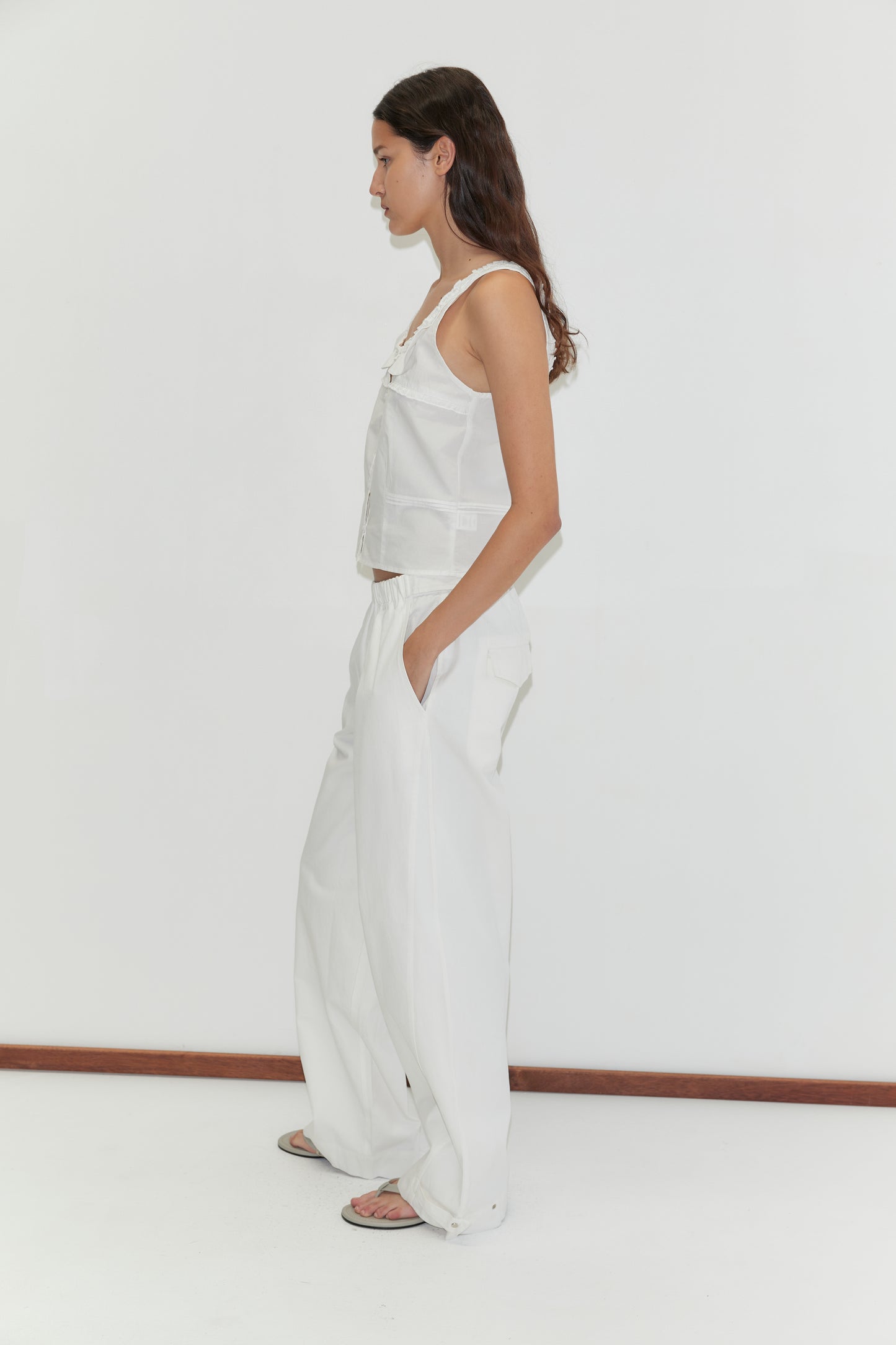 A woman with long dark hair stands in profile against a white background, wearing the Frills Top - White, loose white pants, and sandals. Her hands are in her pockets as she looks straight ahead.
