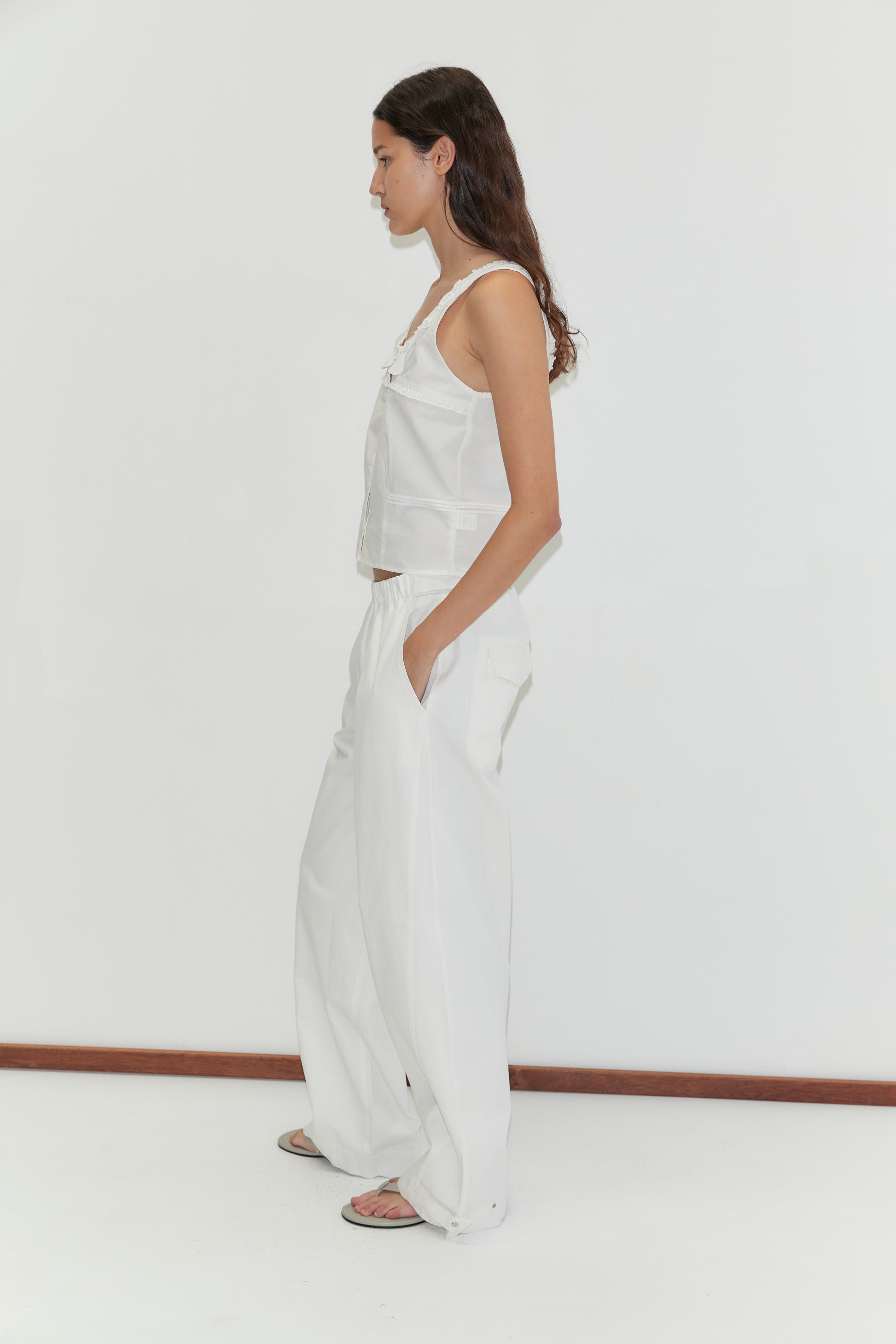 A woman with long dark hair stands in profile against a white background, wearing the Frills Top - White, loose white pants, and sandals. Her hands are in her pockets as she looks straight ahead.