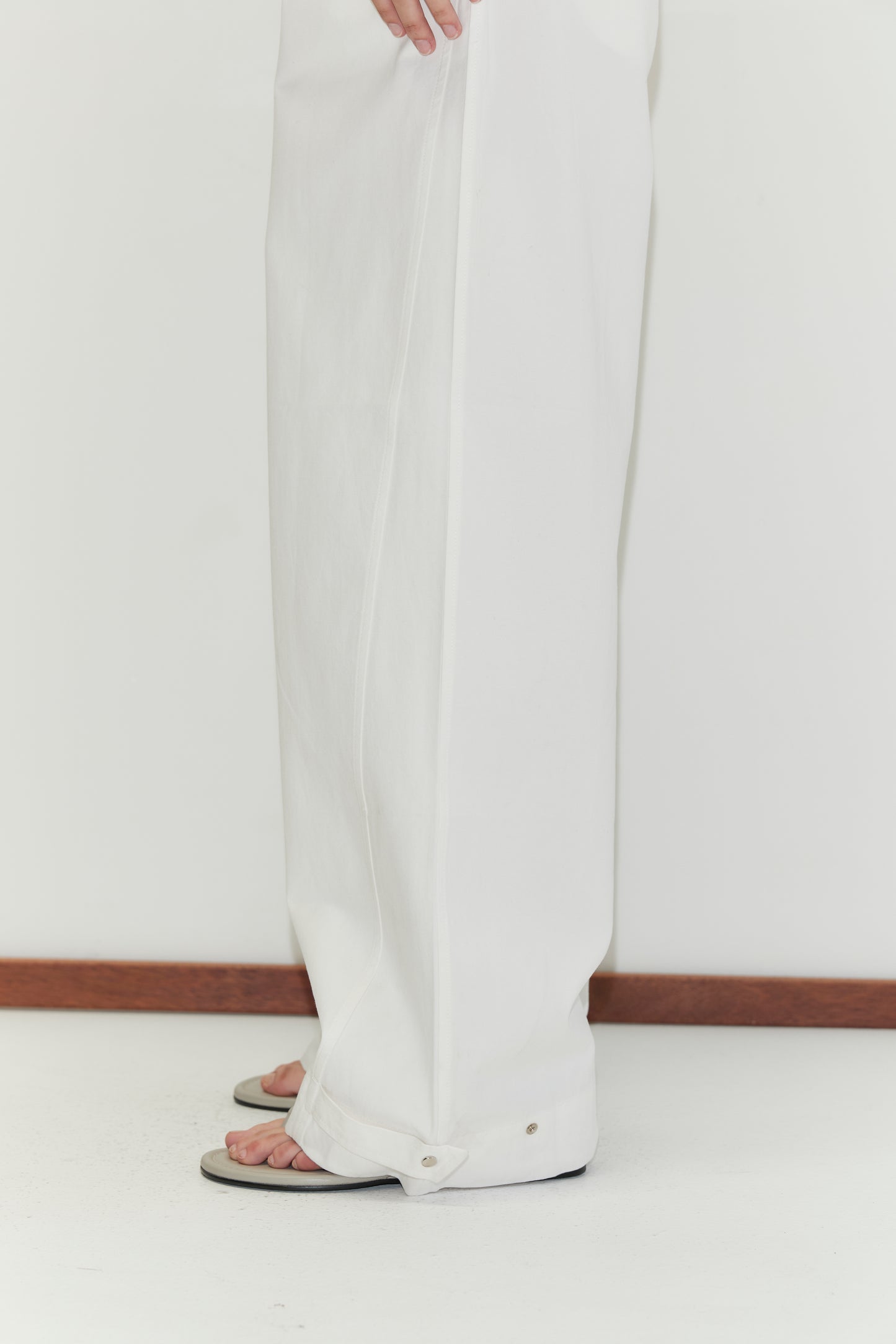 A person wearing Snap Tab Pant - White by Deiji Studios and sandals stands on a white floor near a wooden baseboard, with only their lower legs and feet visible.