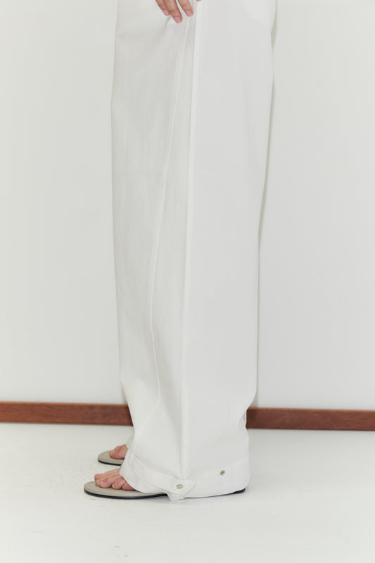 A person wearing Snap Tab Pant - White by Deiji Studios and sandals stands on a white floor near a wooden baseboard, with only their lower legs and feet visible.
