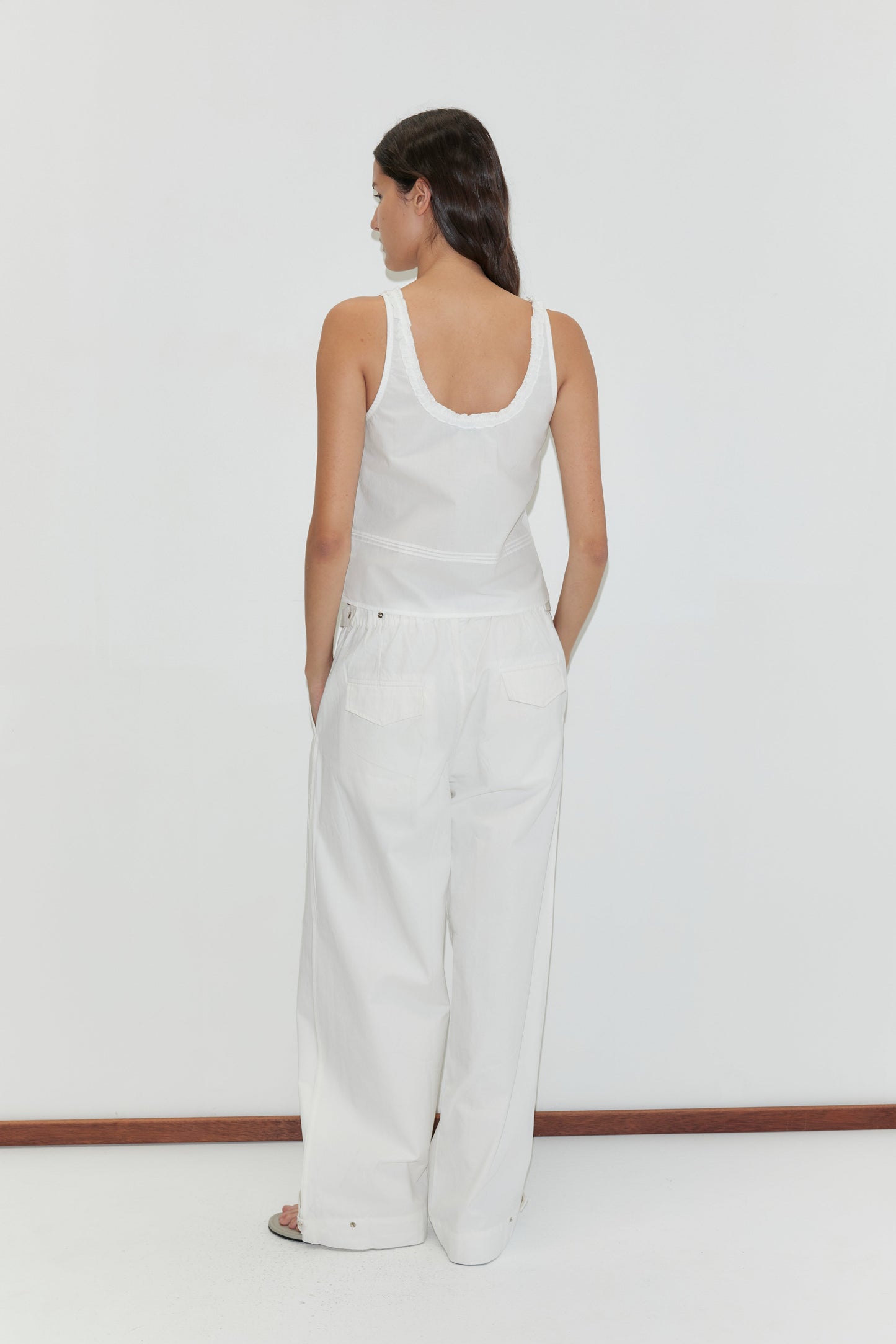 A woman with long dark hair faces away, wearing the Frills Top - White by Deiji Studios and wide-leg white pants, standing before a plain white background.