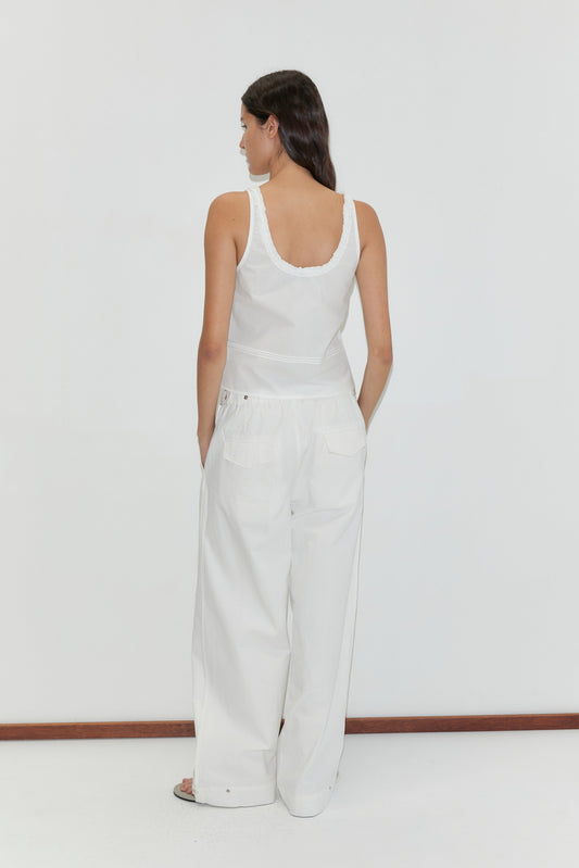 A woman with long dark hair faces away, wearing the Frills Top - White by Deiji Studios and wide-leg white pants, standing before a plain white background.
