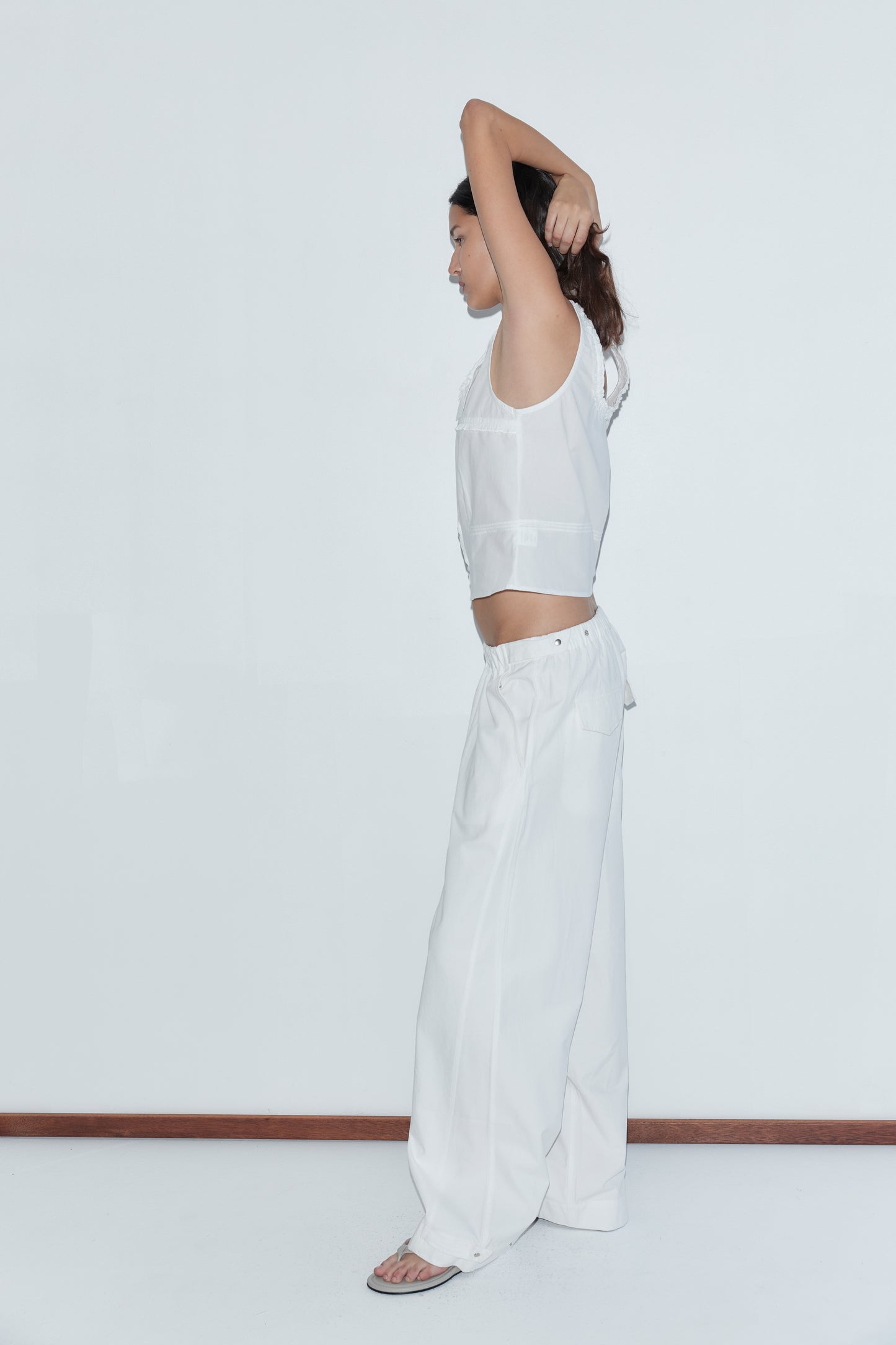 Wearing the Snap Tab Pant - White, a person stands in profile against a plain white wall, dressed in loose white pants and a sleeveless top, with one arm raised and the other bent behind their head.