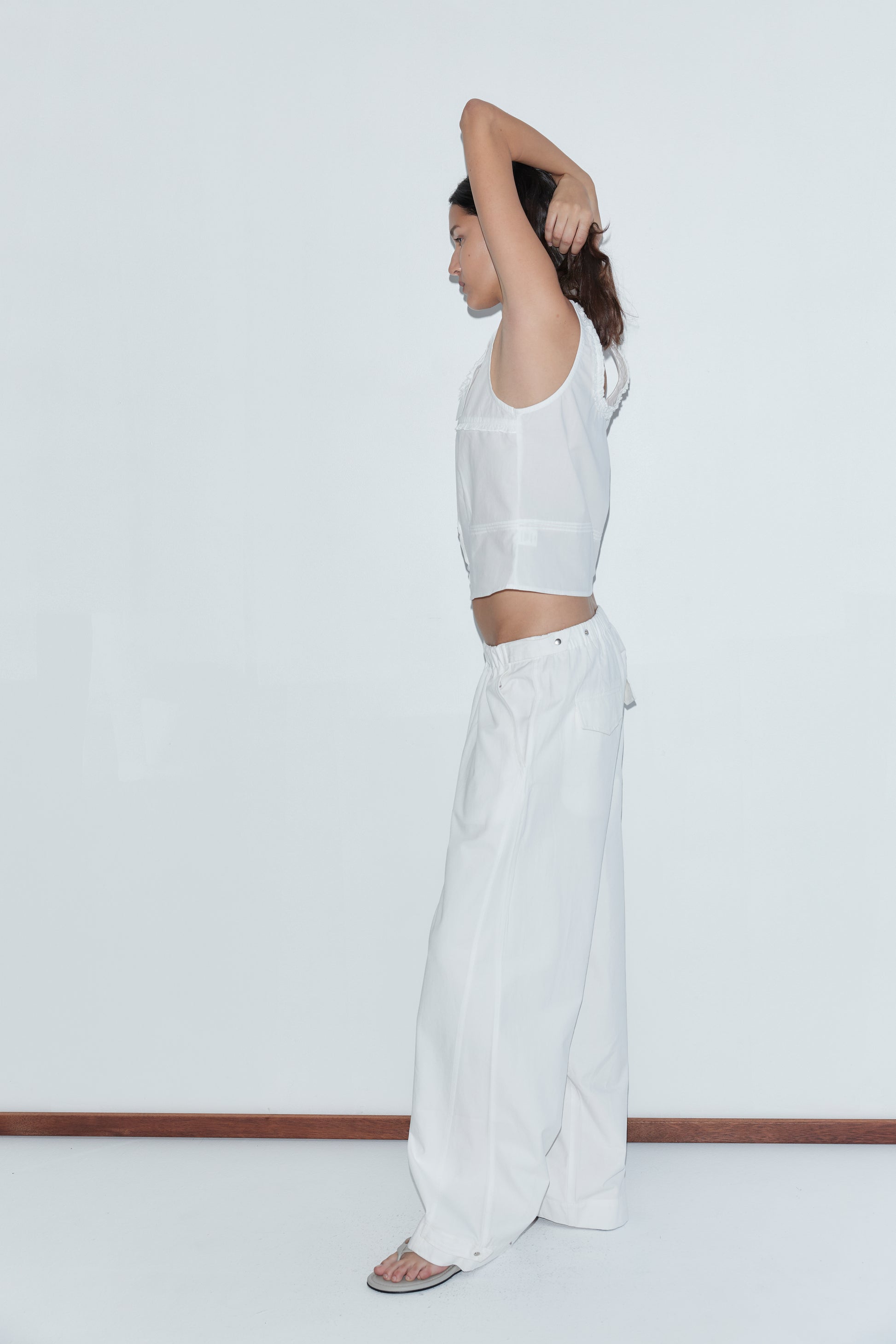 Wearing the Snap Tab Pant - White, a person stands in profile against a plain white wall, dressed in loose white pants and a sleeveless top, with one arm raised and the other bent behind their head.