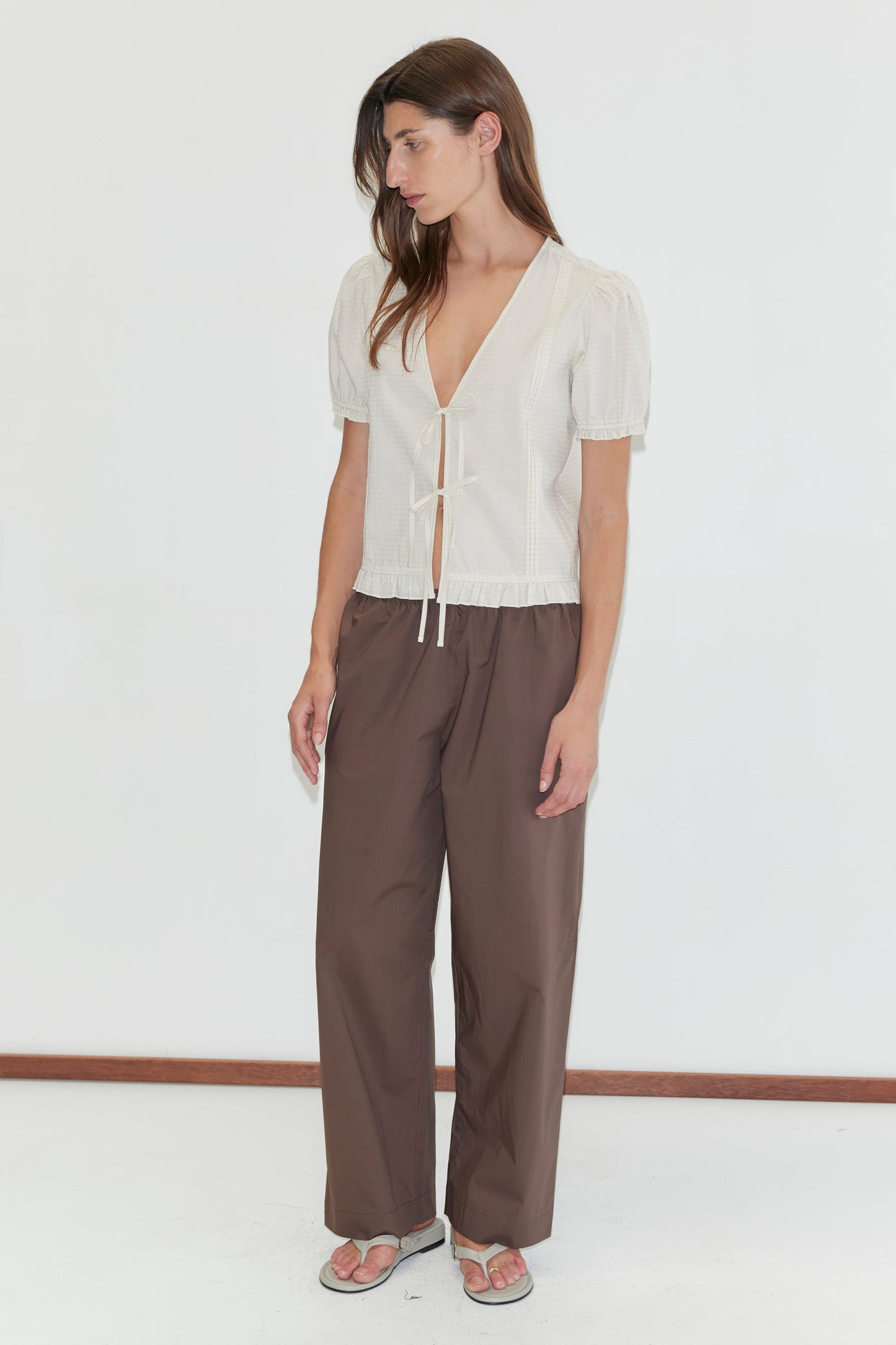 A woman stands against a plain white background wearing the Deiji Studios Pintuck Top in Bone Check, featuring short sleeves, a deep V-neck with tie front, loose brown trousers, and sandals. She gazes slightly downward and to the side.