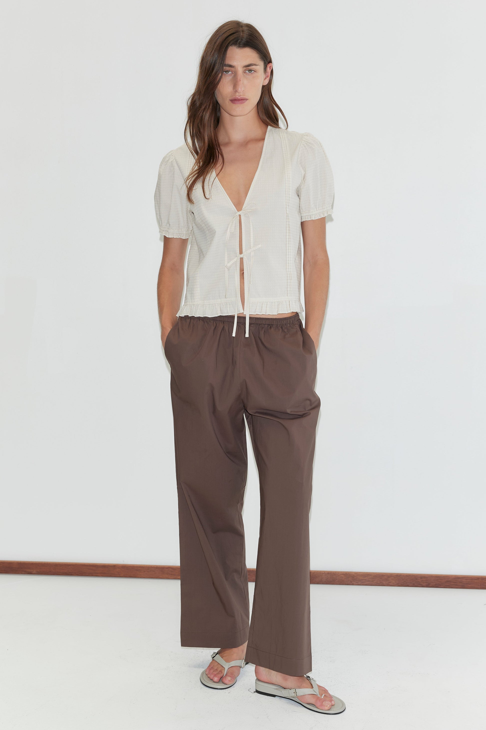 A woman stands against a white background wearing the Deiji Studios Pintuck Top - Bone Check, paired with loose brown pants and sandals. Her hands are in her pockets as she faces the camera.