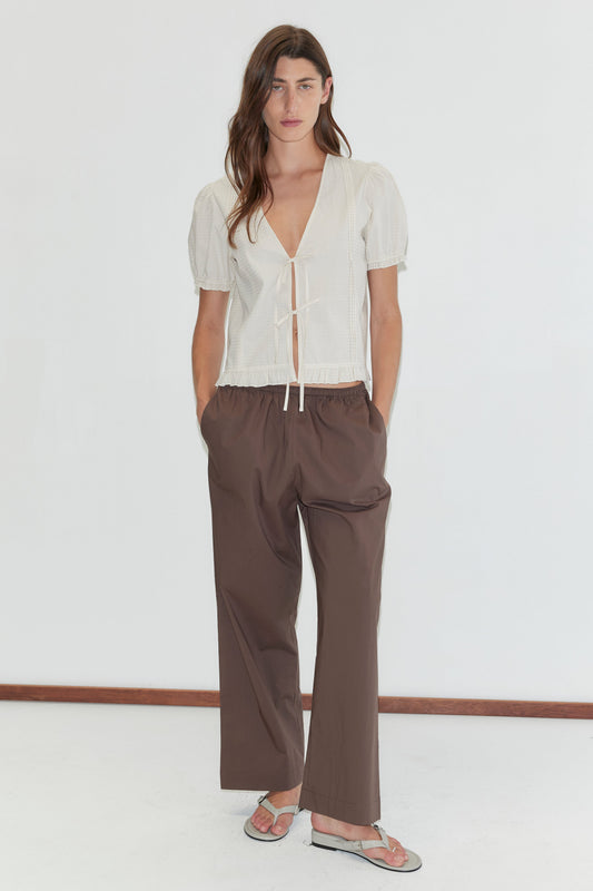 A woman stands against a white background wearing the Deiji Studios Pintuck Top - Bone Check, paired with loose brown pants and sandals. Her hands are in her pockets as she faces the camera.