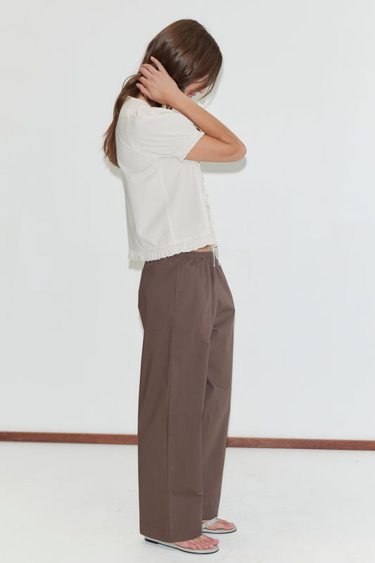 A woman in profile wears the Deiji Studios Pintuck Top - Bone Check, featuring a fringe hem, with wide-leg brown trousers and silver sandals, standing against a white background.