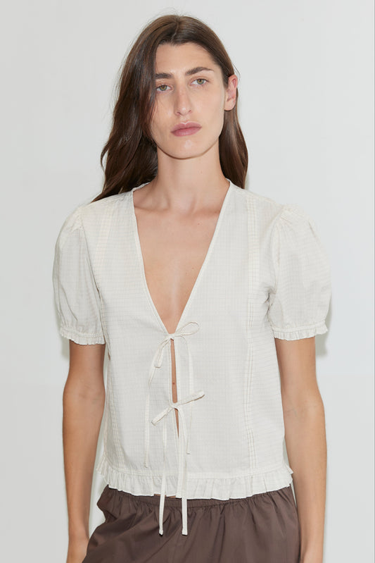 A person with long brown hair wears the Deiji Studios Pintuck Top in Bone Check, featuring short blouson sleeves and a deep V-neck with two bows, paired with brown bottoms, standing against a plain light background.