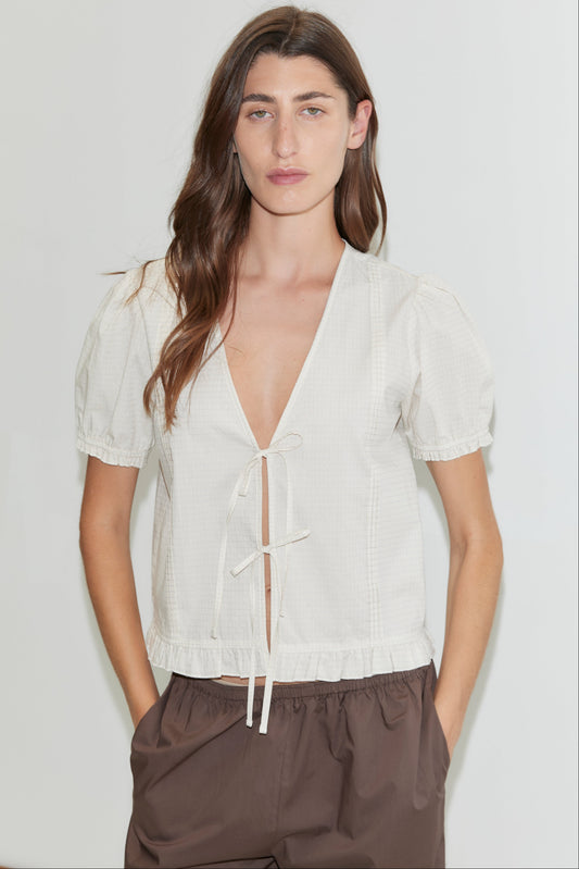 A woman stands against a plain background wearing the Pintuck Top - Bone Check by Deiji Studios with front ties and loose brown trousers. She has long, wavy brown hair, a neutral expression, and her hands in her pockets.