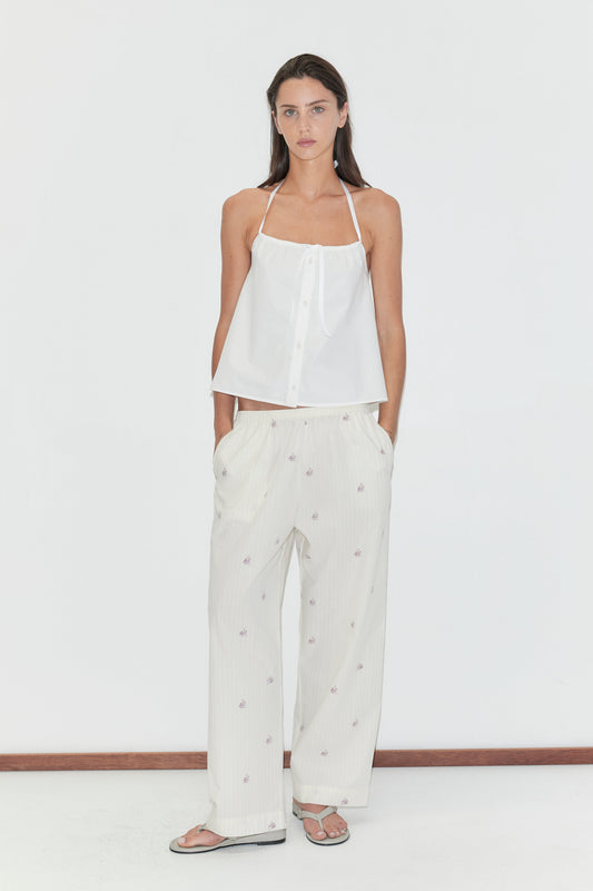 A woman stands against a white background, wearing a white halter top and Deiji Studios’ Ease Trouser - Floral Stripe, featuring a subtle floral stripe pattern. She pairs the look with flip-flops, her hands casually in her pockets, and has long, straight dark hair.