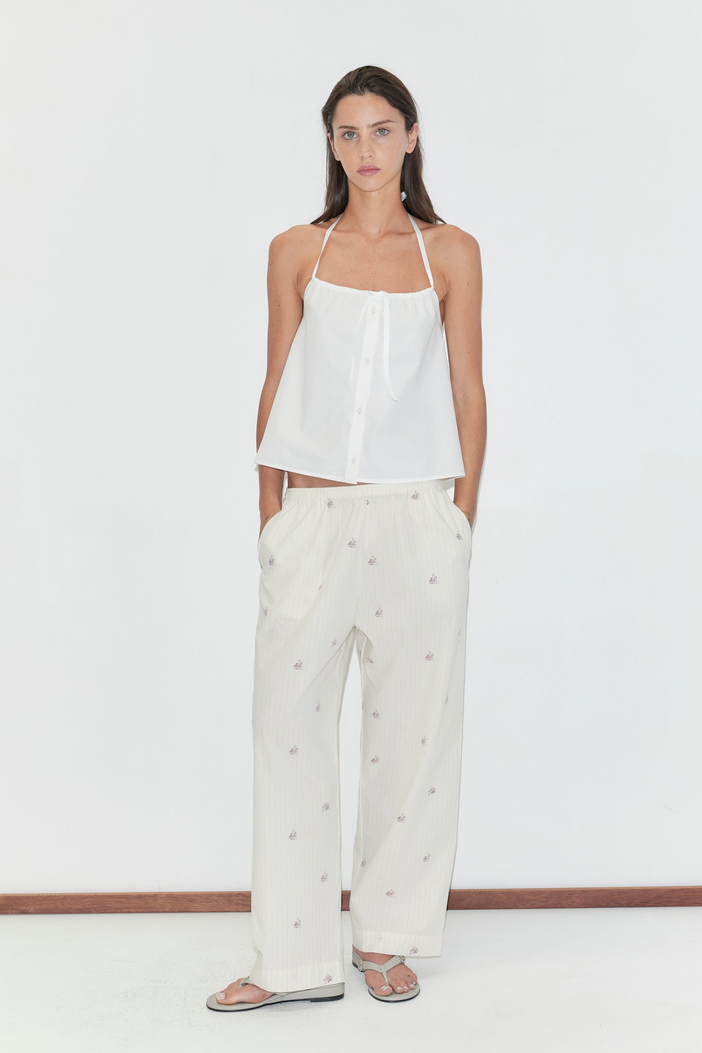 A woman with long dark hair stands against a plain white background wearing the Simple Tie Top - White by Deiji Studios, a white halter top with a gathered neckline and front tie, with cream floral trousers and flip-flops.