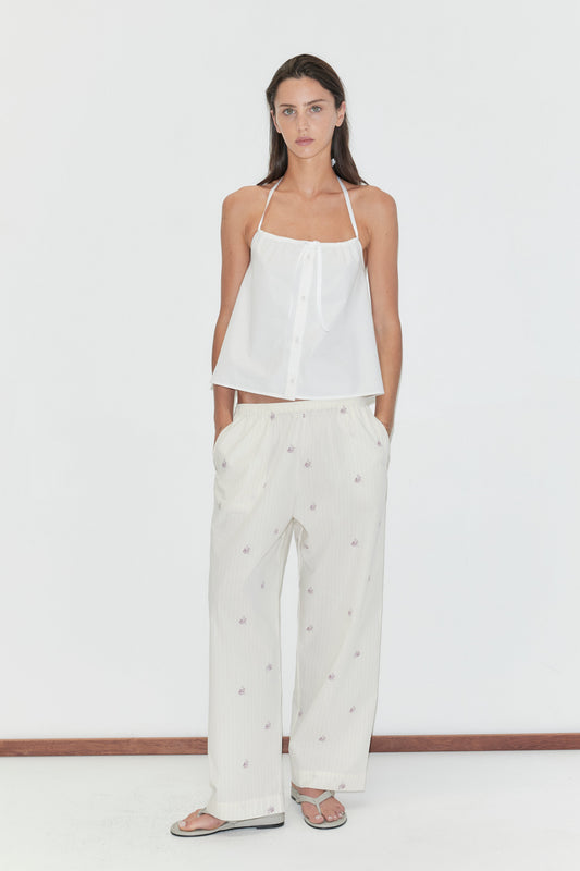 A woman with long dark hair stands against a plain white background wearing the Simple Tie Top - White by Deiji Studios, a white halter top with a gathered neckline and front tie, with cream floral trousers and flip-flops.
