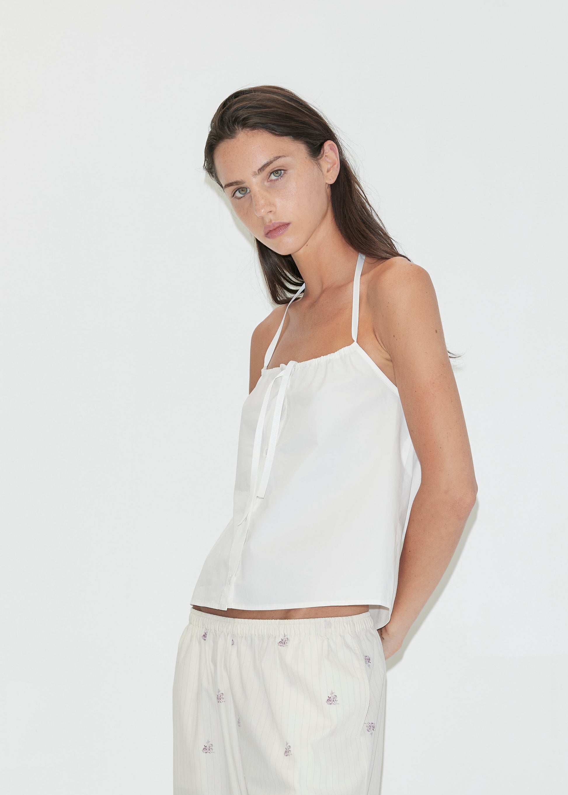 A woman with long dark hair stands against a plain white background wearing the Simple Tie Top - White by Deiji Studios, a white halter top with a gathered neckline and drawstring front tie, with cream floral trousers.