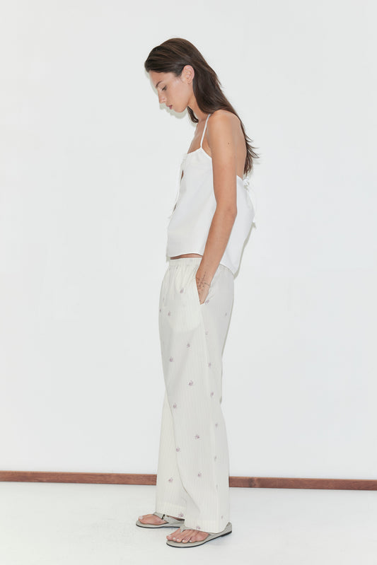 A woman stands sideways against a plain white background, looking down with her hands in the pockets of her light-colored Ease Trouser - Floral Stripe and wearing a white sleeveless top and sandals—a relaxed look by Deiji Studios.