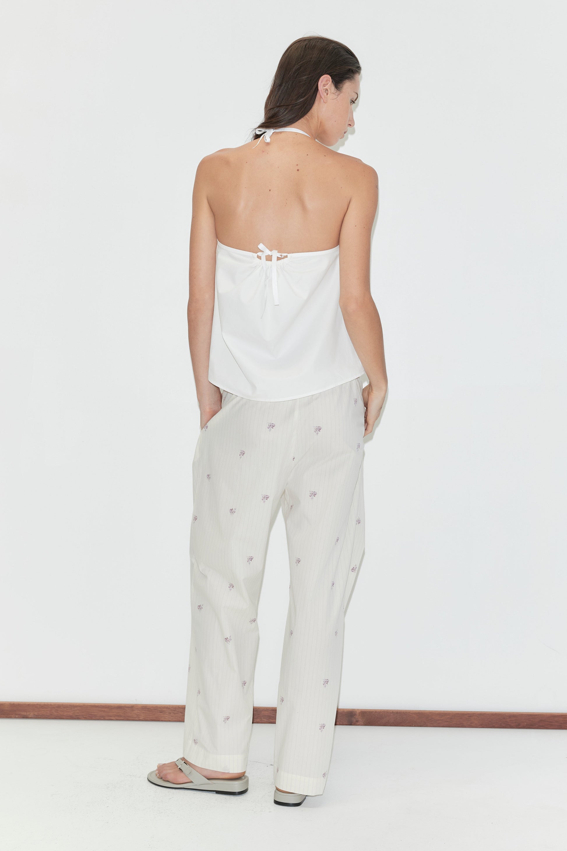 A woman with long dark hair stands with her back to the camera against a plain white background wearing the Simple Tie Top - White by Deiji Studios, a white halter top with an open back and tie fastening, with cream floral trousers.