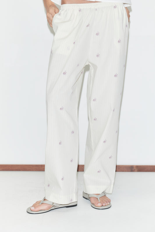 A person is wearing the Ease Trouser - Floral Stripe from Deiji Studios, featuring loose white pajama pants with a subtle pink floral stripe pattern, standing on a white floor with sandals; their upper body is partially out of frame.