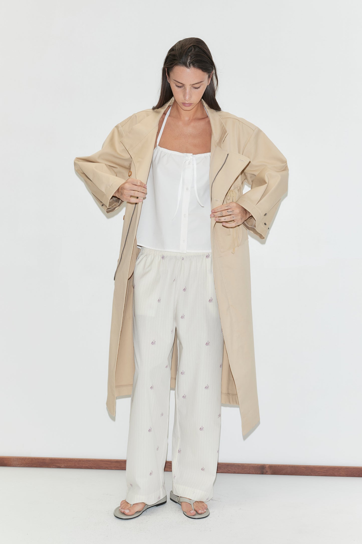 A woman with long dark hair stands against a plain white background looking down, wearing the Simple Tie Top - White by Deiji Studios, a white halter top with a front tie, under an open beige trench coat with cream floral trousers and flip-flops.