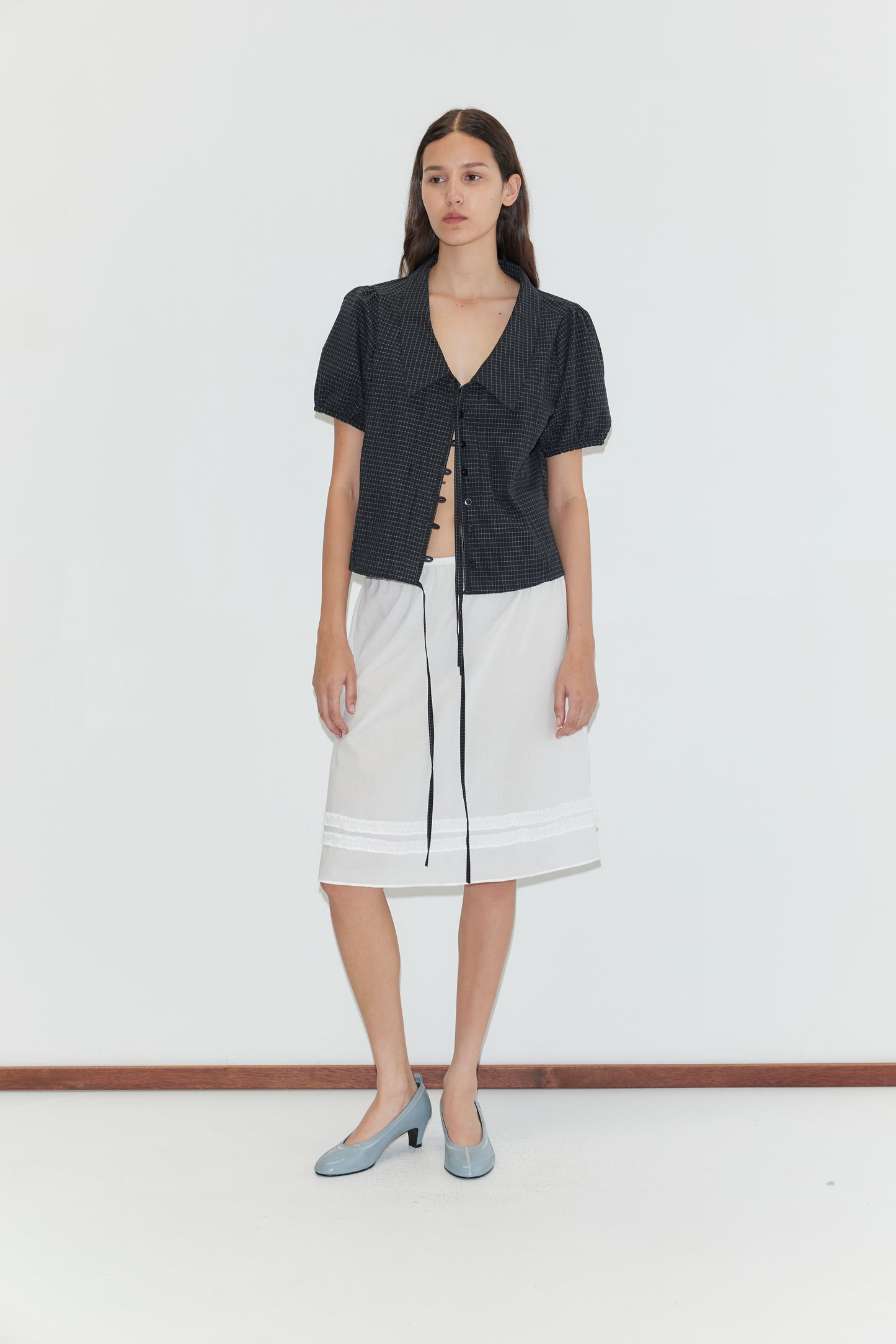 A woman with long brown hair stands against a white background, wearing a short-sleeved dark blouse with a tie front, the Daytime Skirt - White, and light blue low-heeled shoes.