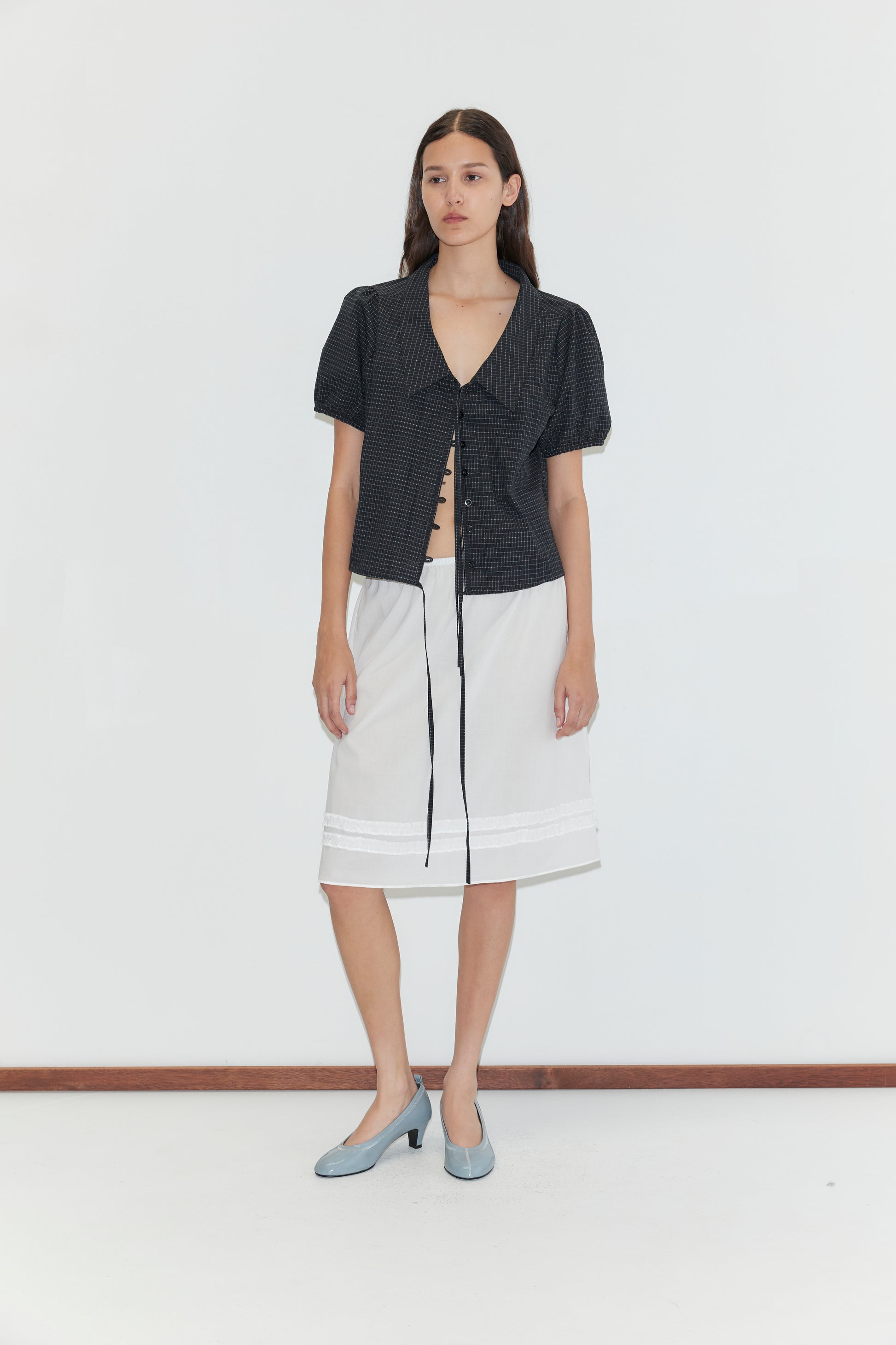 A woman with long brown hair stands against a white background, wearing a short-sleeved dark blouse with a tie front, the Daytime Skirt - White, and light blue low-heeled shoes.