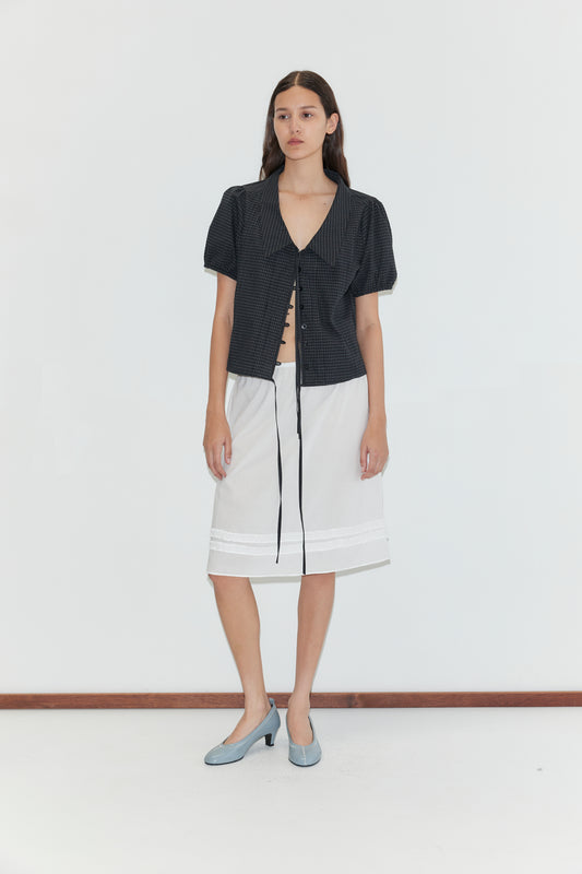 A woman with long brown hair stands against a white background, wearing a short-sleeved dark blouse with a tie front, the Daytime Skirt - White, and light blue low-heeled shoes.