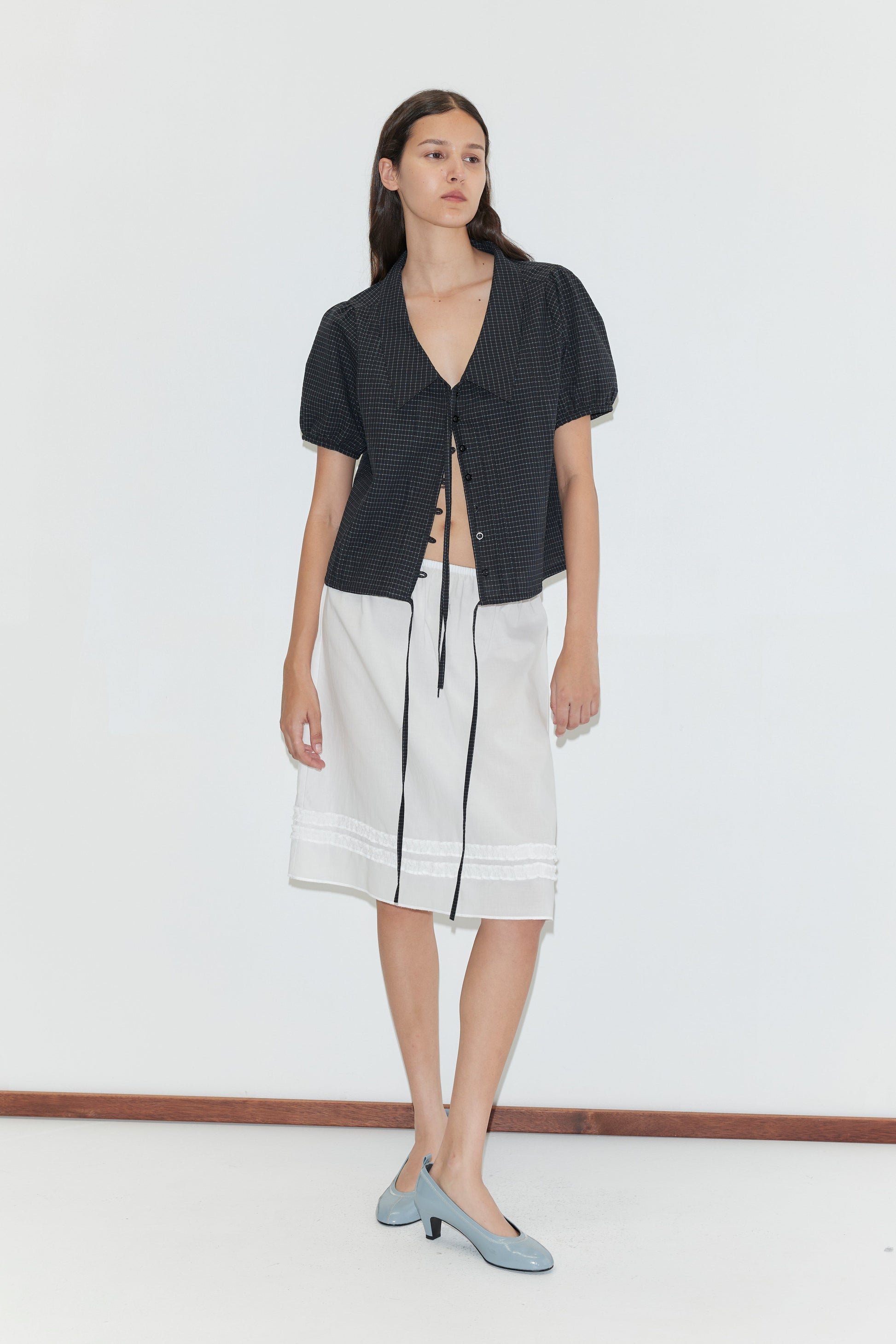 A woman with long brown hair and a neutral expression stands against a plain white background wearing the Fine Collar Top - Mono Grid, a white knee-length skirt with lace trim, and light blue heels.