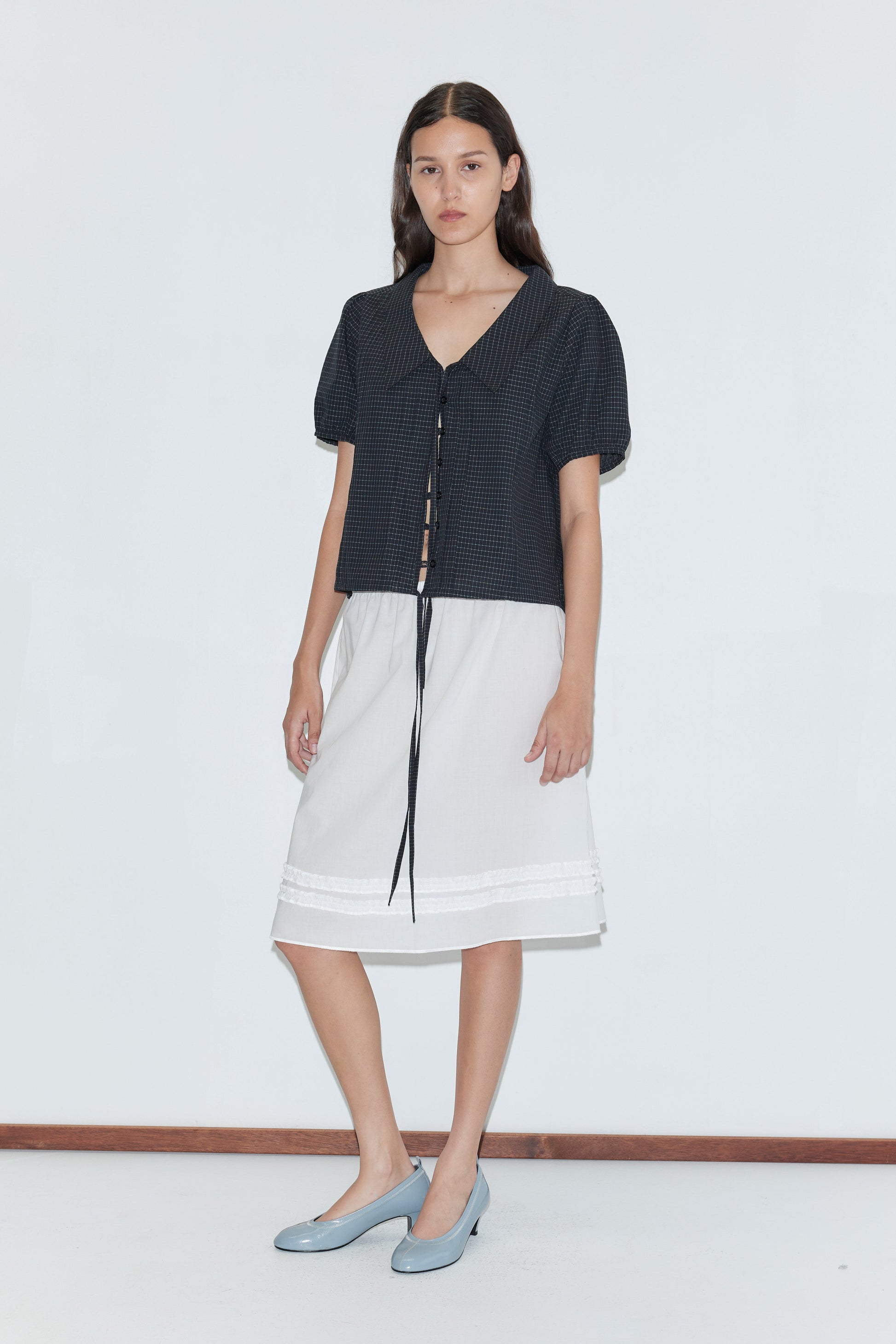 A woman with long brown hair stands against a white wall, wearing the Fine Collar Top - Mono Grid and a knee-length white skirt with lace trim. She completes the look with light blue low-heeled shoes.
