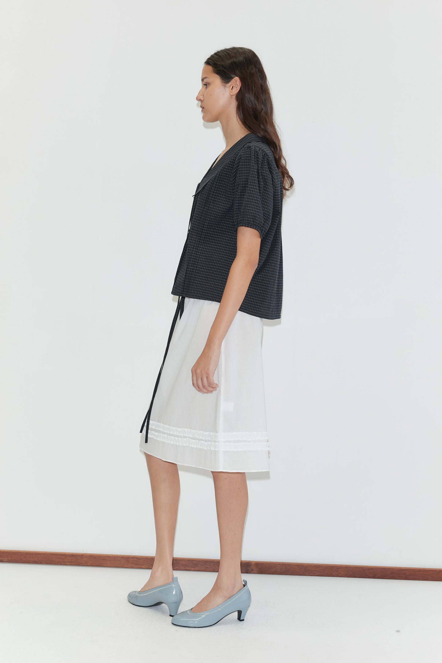 A woman stands in profile against a white wall, wearing the Fine Collar Top - Mono Grid, a dark checkered cotton poplin collared v-neck, with a white lace-trim skirt and light blue heels—evoking Deiji Studios' effortless style.