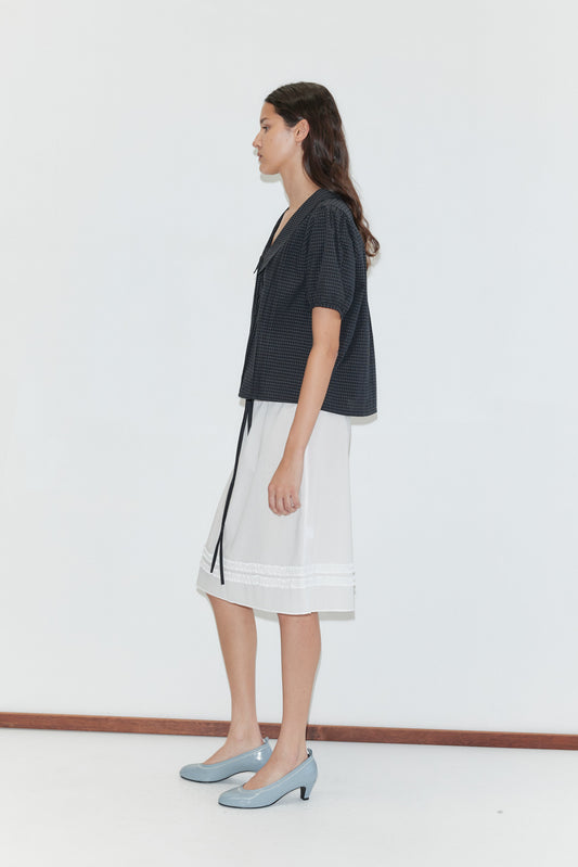 A woman with long dark hair, in a loose dark short-sleeve blouse, the Daytime Skirt - White, and light blue heels stands in profile against a plain white background.