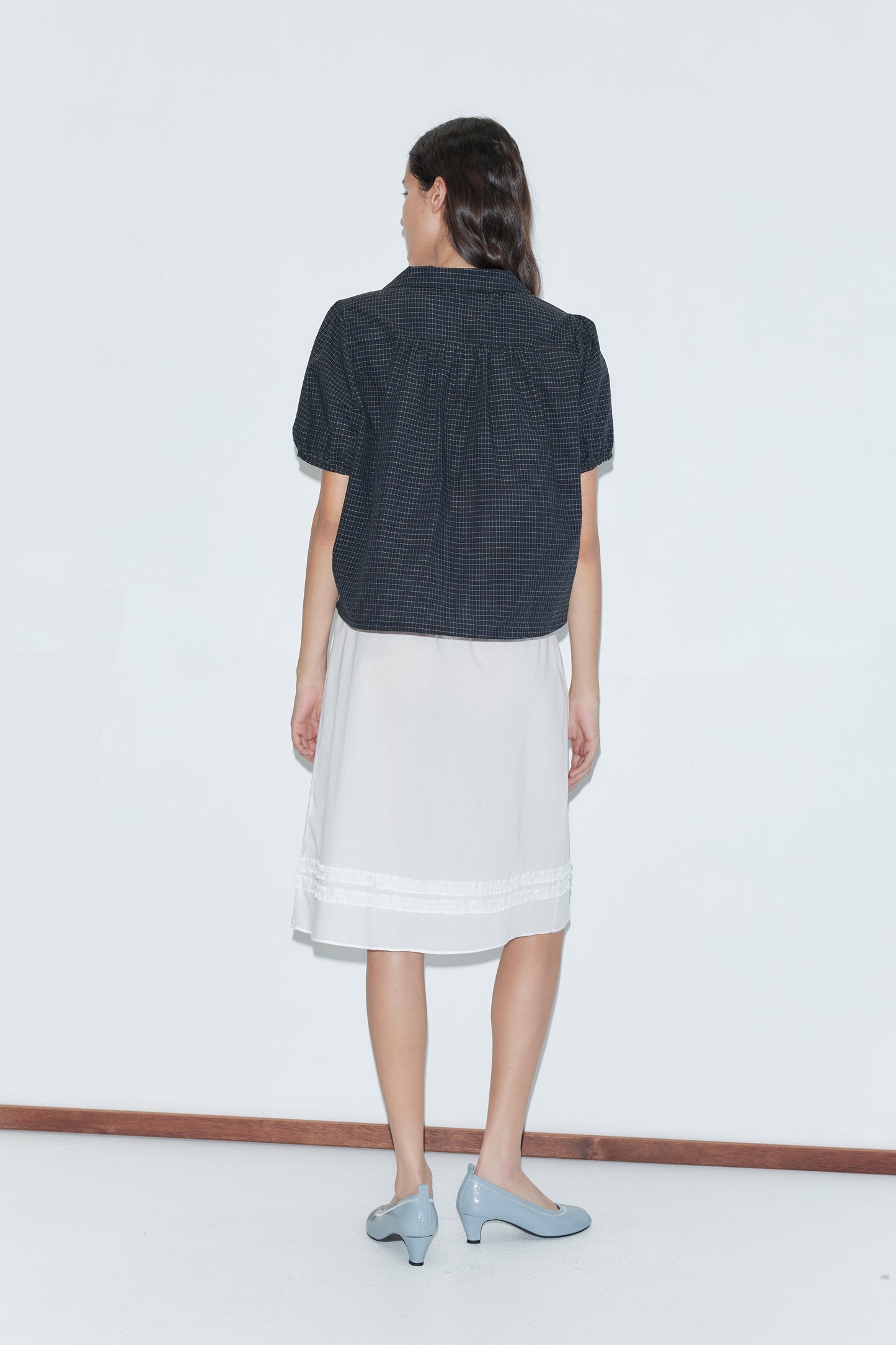A woman faces away from the camera, wearing a short-sleeved dark checkered shirt over the Daytime Skirt - White and light blue heeled shoes, set against a plain white background.