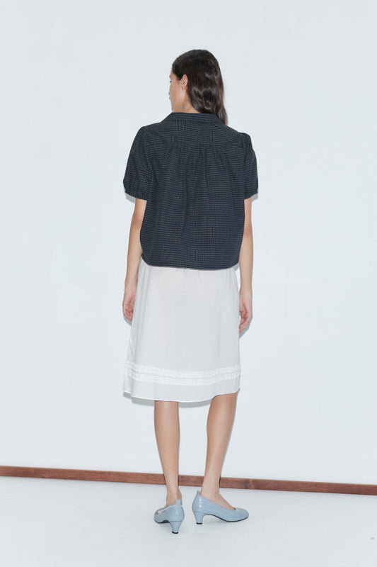 A woman faces away from the camera, wearing a short-sleeved dark checkered shirt over the Daytime Skirt - White and light blue heeled shoes, set against a plain white background.