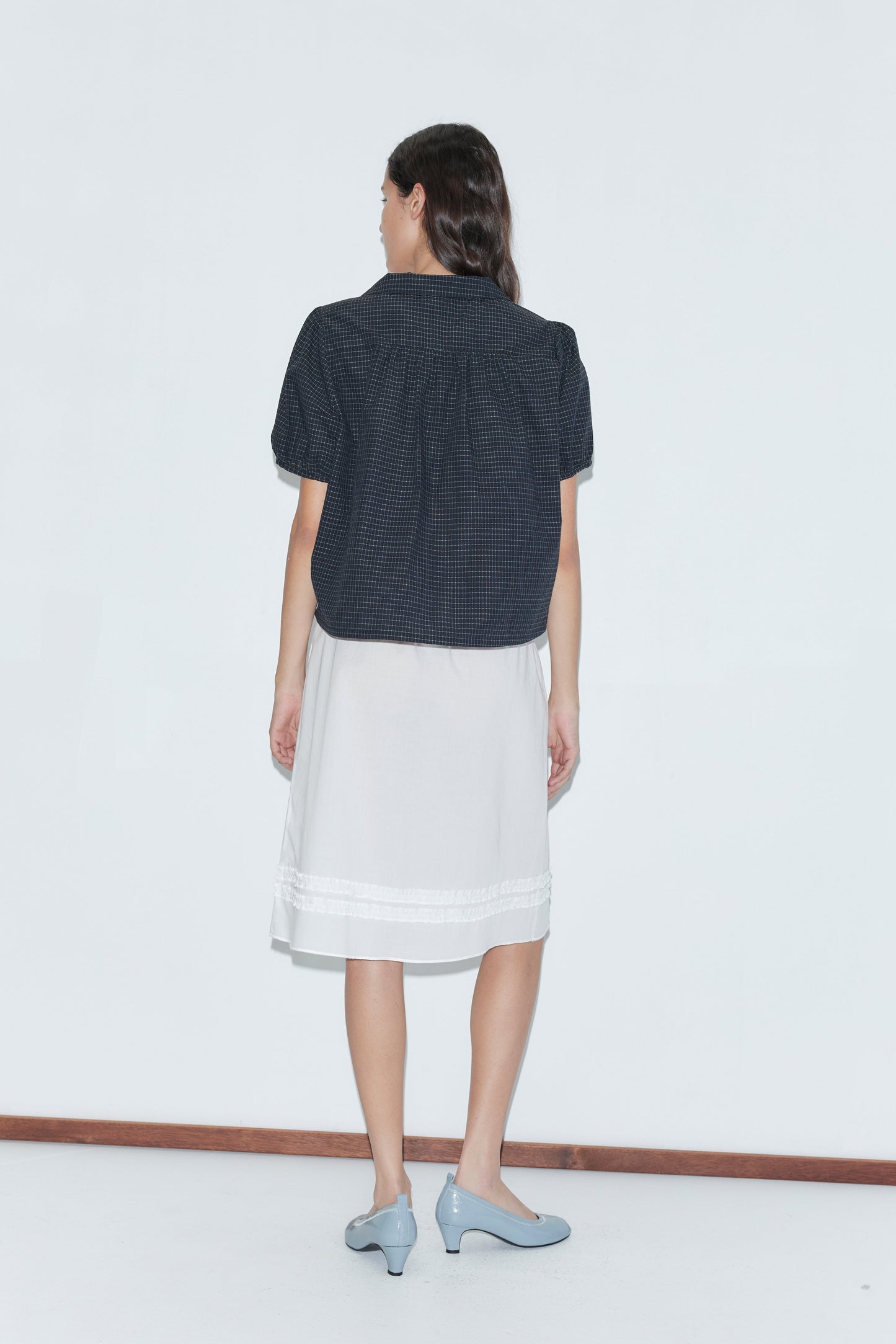 A woman faces away, wearing the Fine Collar Top - Mono Grid by Deiji Studios, a knee-length white skirt, and light blue heels, set against a plain white background.