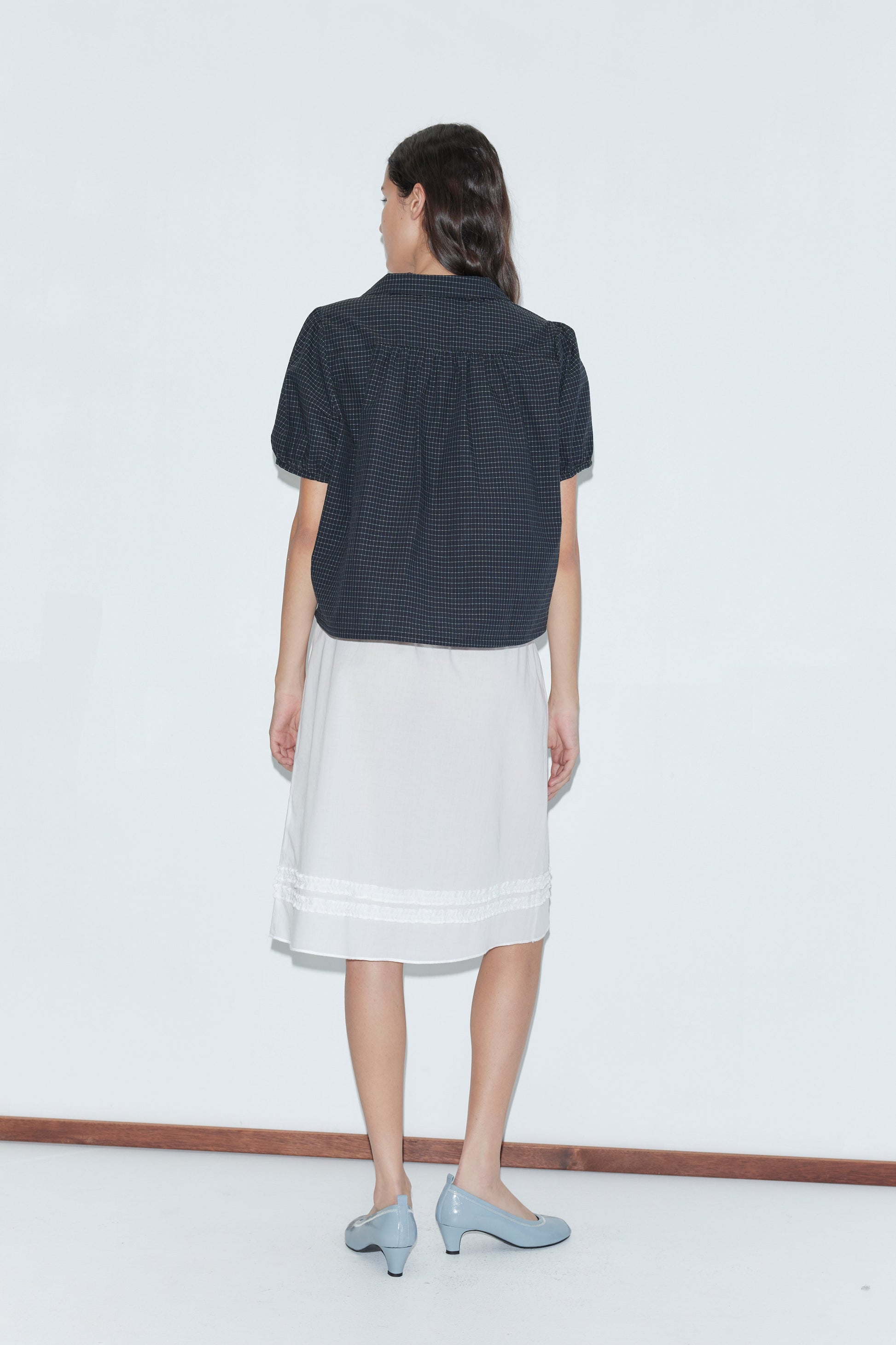 A woman faces away, wearing the Fine Collar Top - Mono Grid by Deiji Studios, a knee-length white skirt, and light blue heels, set against a plain white background.
