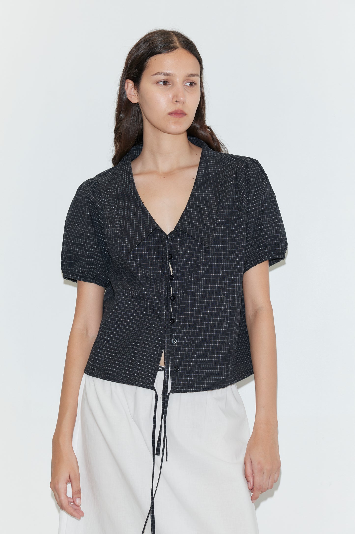 A woman with long brown hair wears the Fine Collar Top - Mono Grid by Deiji Studios, featuring a collared v-neck and short sleeves, paired with a white skirt against a plain white background.